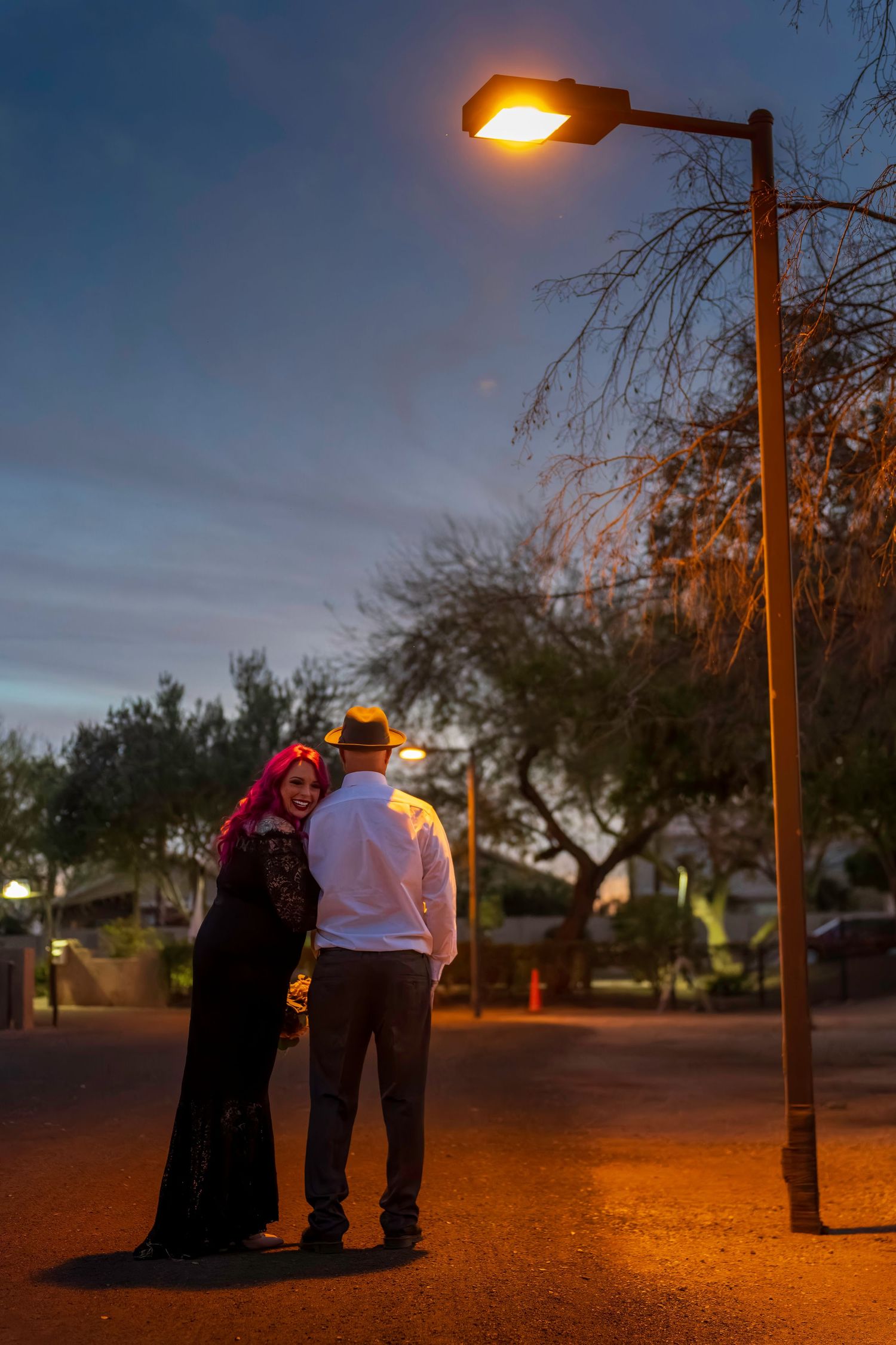 A couple with pink and white hair embrace under a glowing street lamp at twilight in an outdoor setting.