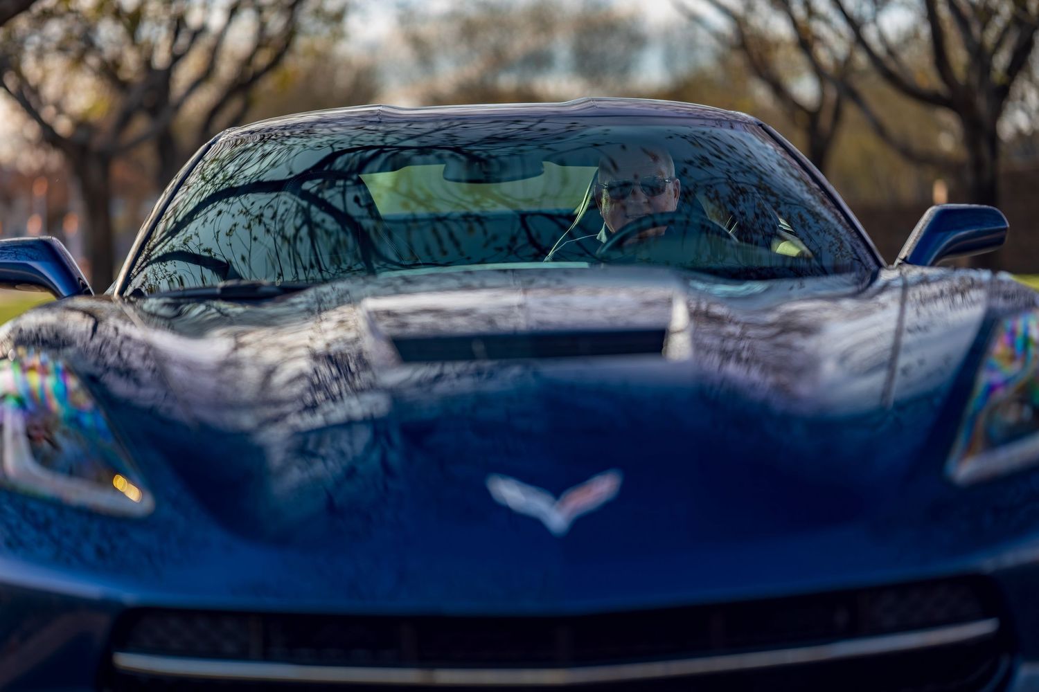 Close-up front view of a dark blue Chevrolet Corvette Stingray with driver visible through windshield.