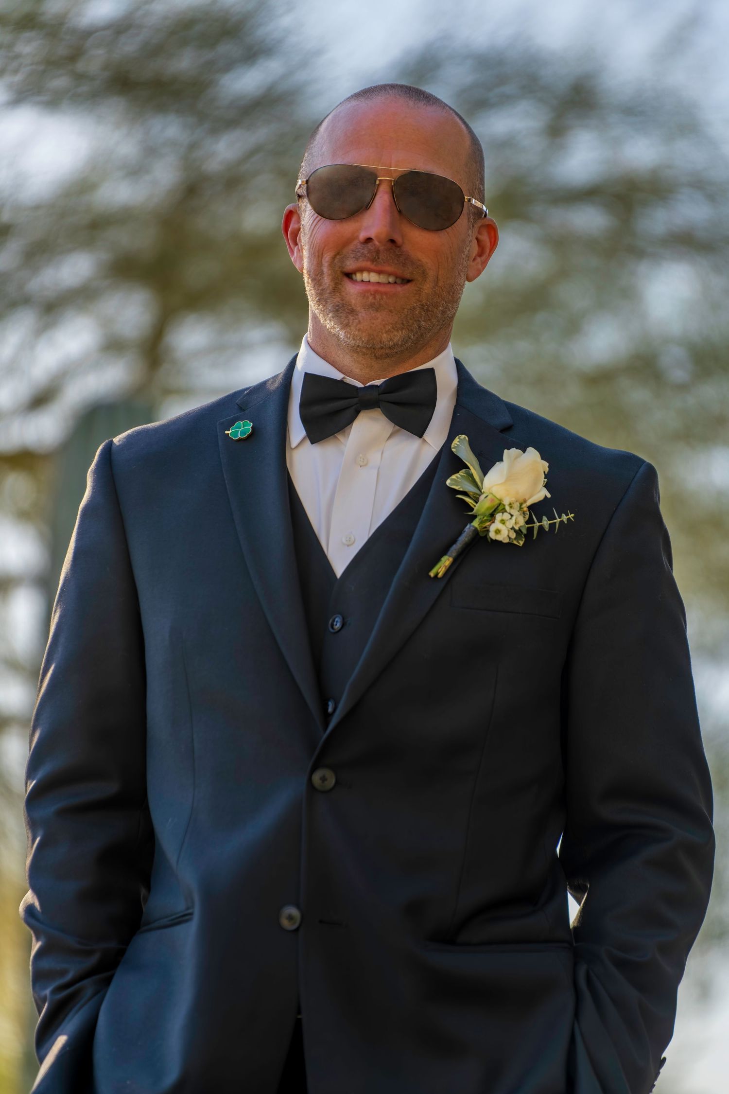 Smiling bald groom in navy tuxedo with bow tie, boutonniere, and aviator sunglasses poses outdoors at wedding.