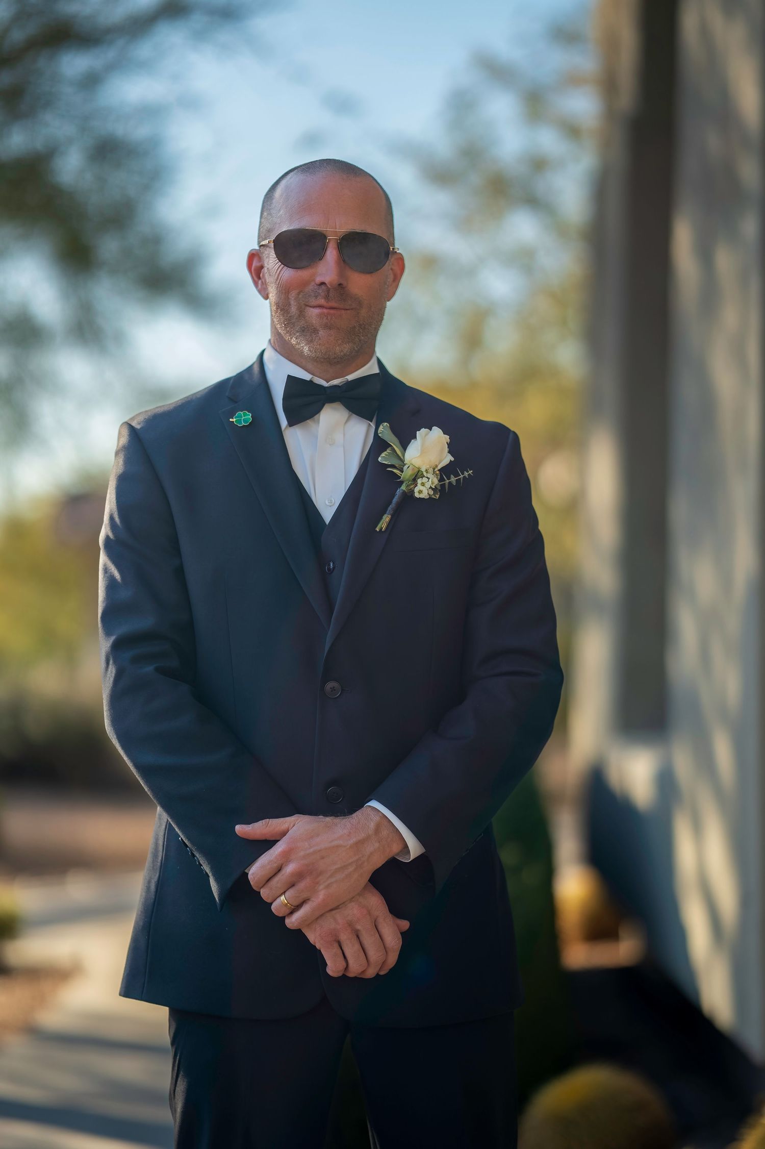 Elegant groom in navy three-piece tuxedo with bow tie and boutonniere wearing sunglasses outdoors.