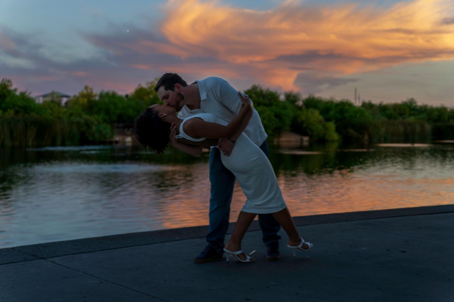 Couple sharing a romantic dip kiss by a lake at sunset with colorful pink and orange clouds in the background.