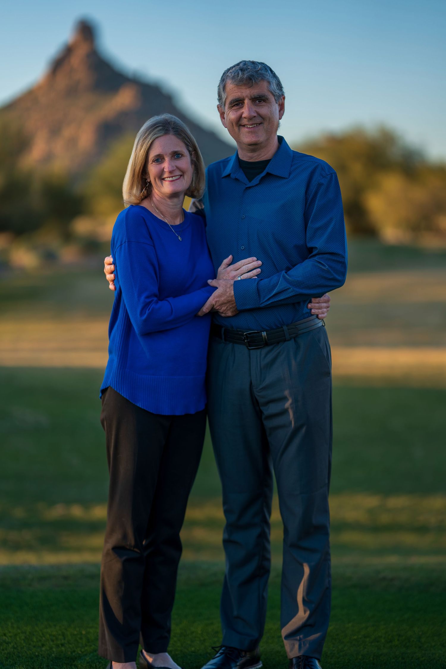 Couple in blue and dark attire posing together on grass with mountain in background.