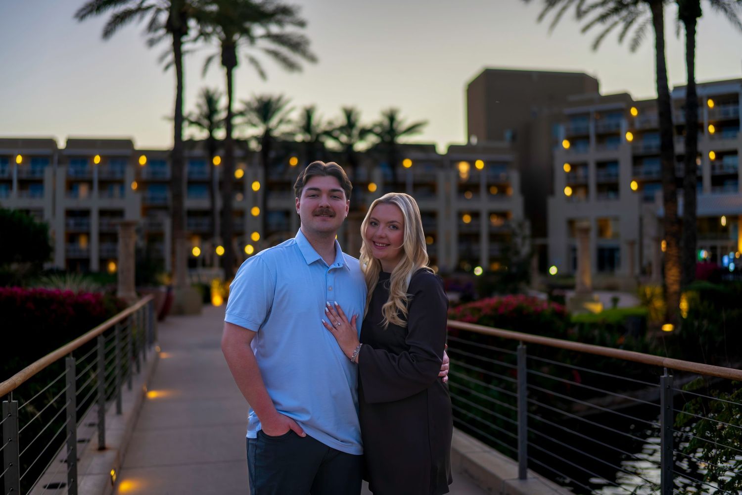 Young couple posing on a bridge at twilight with a lit resort and palm trees behind them.