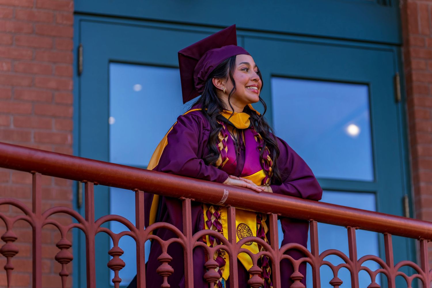 Graduate poses on decorative balcony railing with purple cap and gown against blue evening sky.