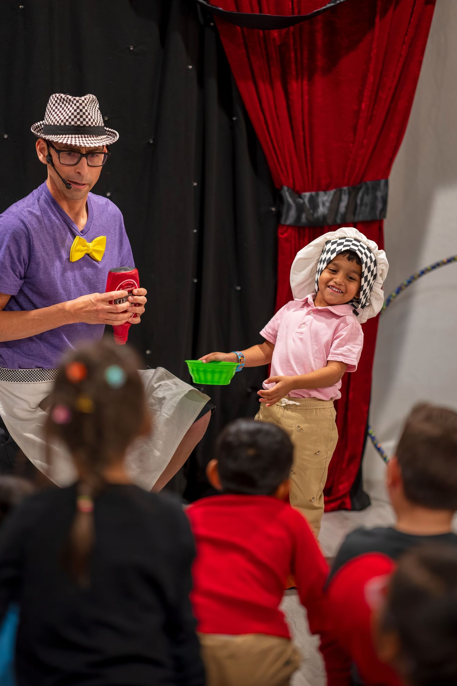 An audience watches attentively during a stage performance with red curtains and theatrical lighting.