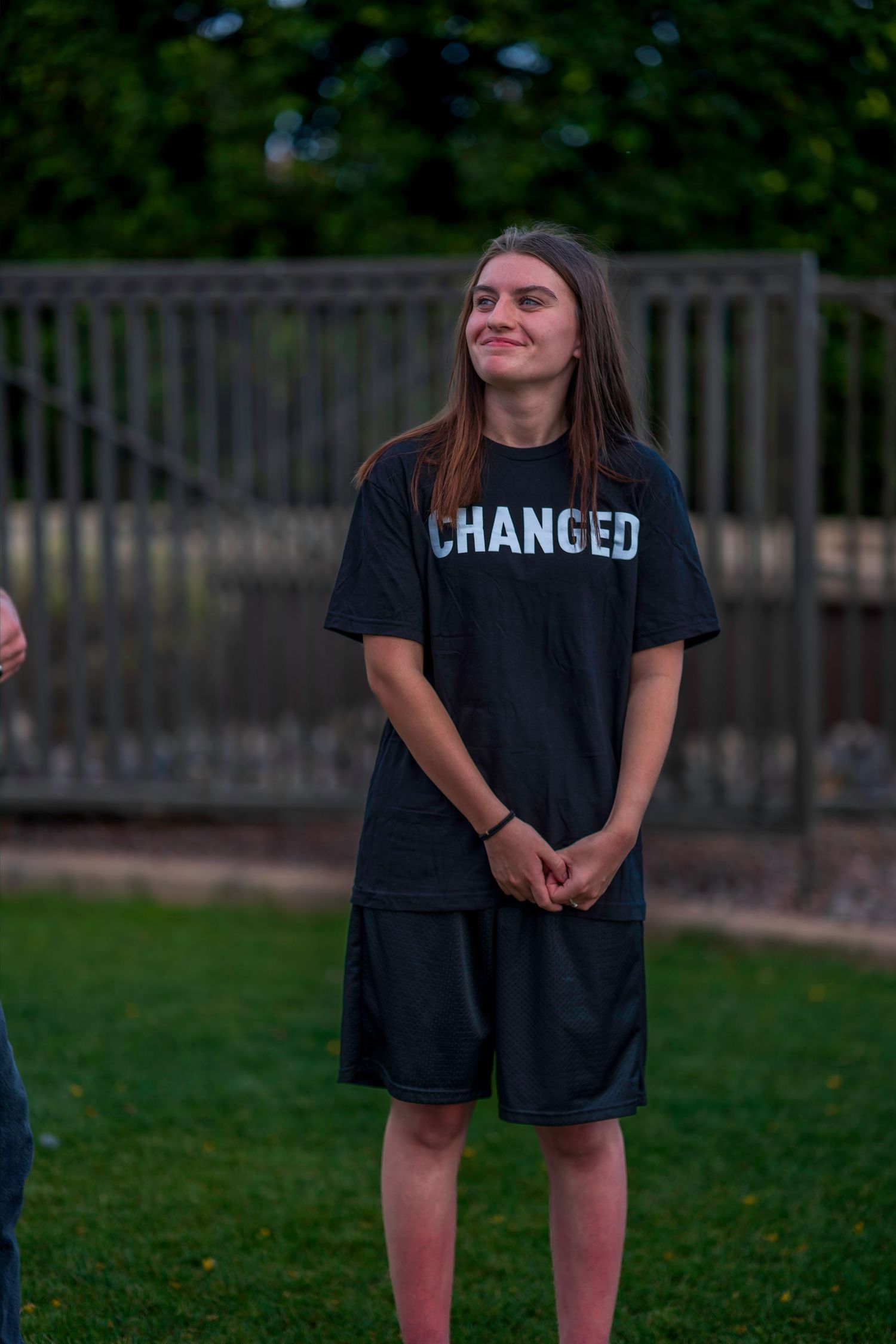 Young woman smiling outdoors wearing a black Changed t-shirt and shorts, standing on green grass.