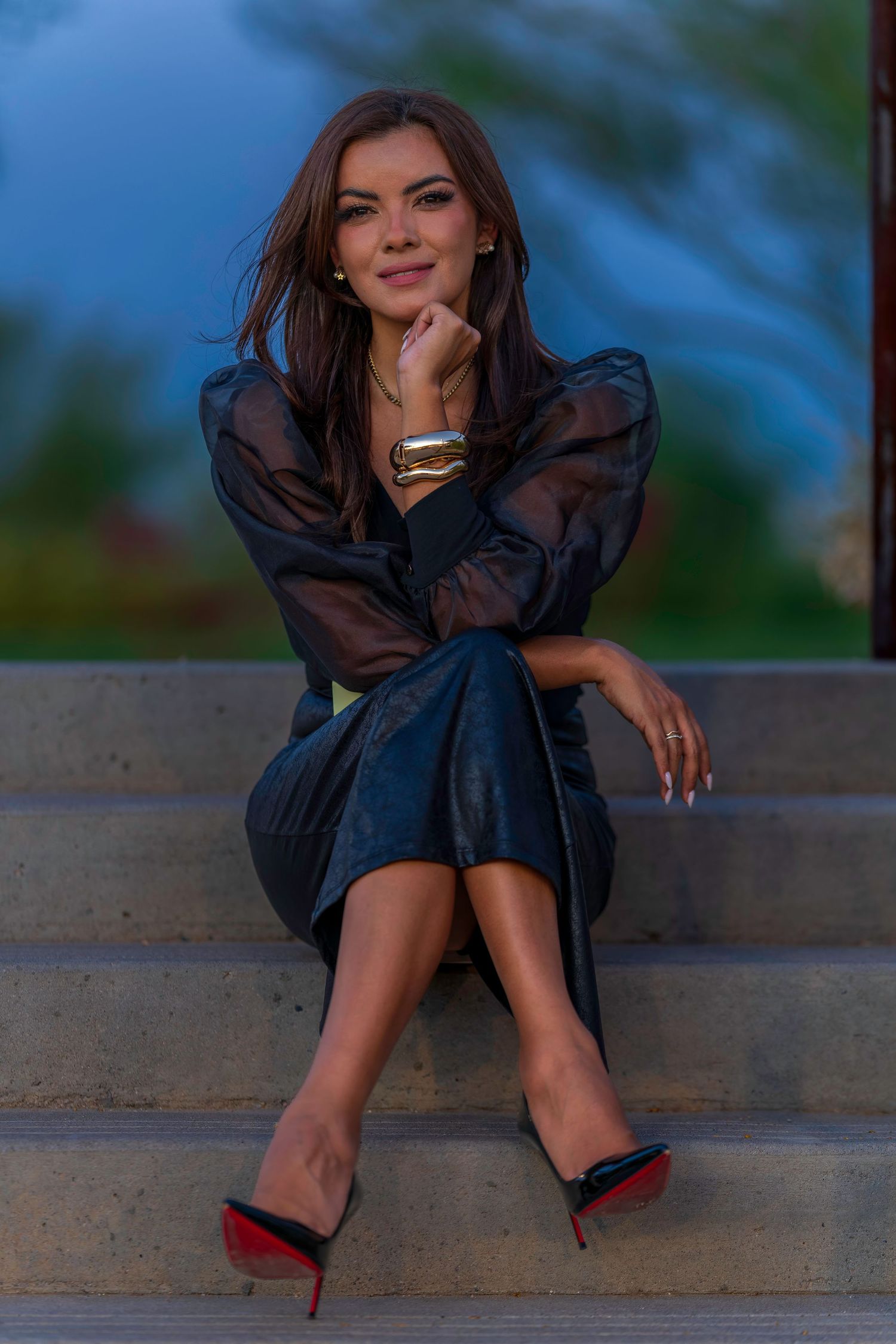 Elegant woman in black sheer blouse and leather skirt with red-sole heels sitting on stone steps outdoors.