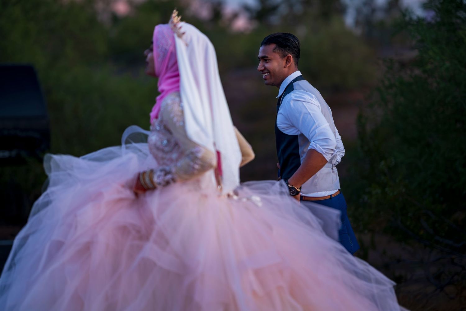 Groom in blue vest helping bride in voluminous pink ball gown wedding dress outdoors at dusk.