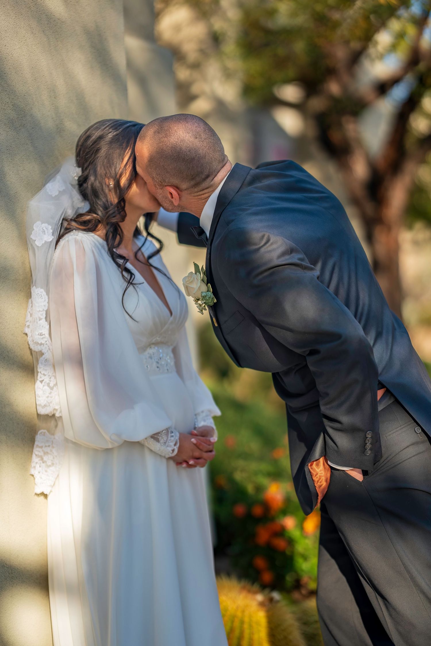 Bride and groom sharing a romantic kiss outdoors, bride in white lace veil and groom in navy suit.