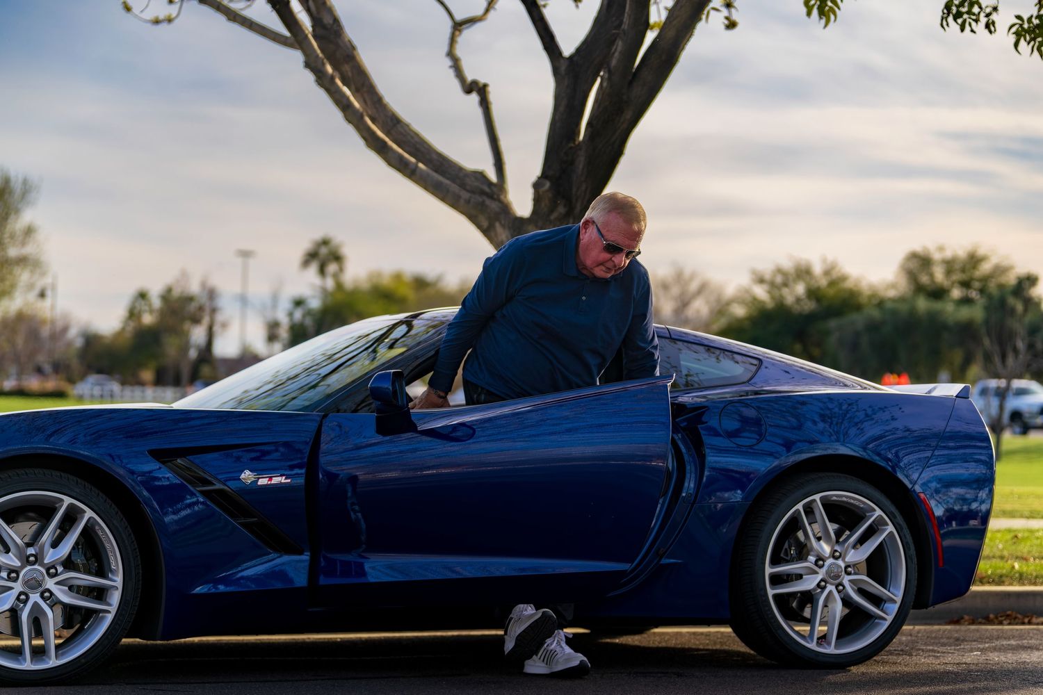 Older man in sunglasses getting into a blue Chevrolet Corvette parked near a large tree at dusk.