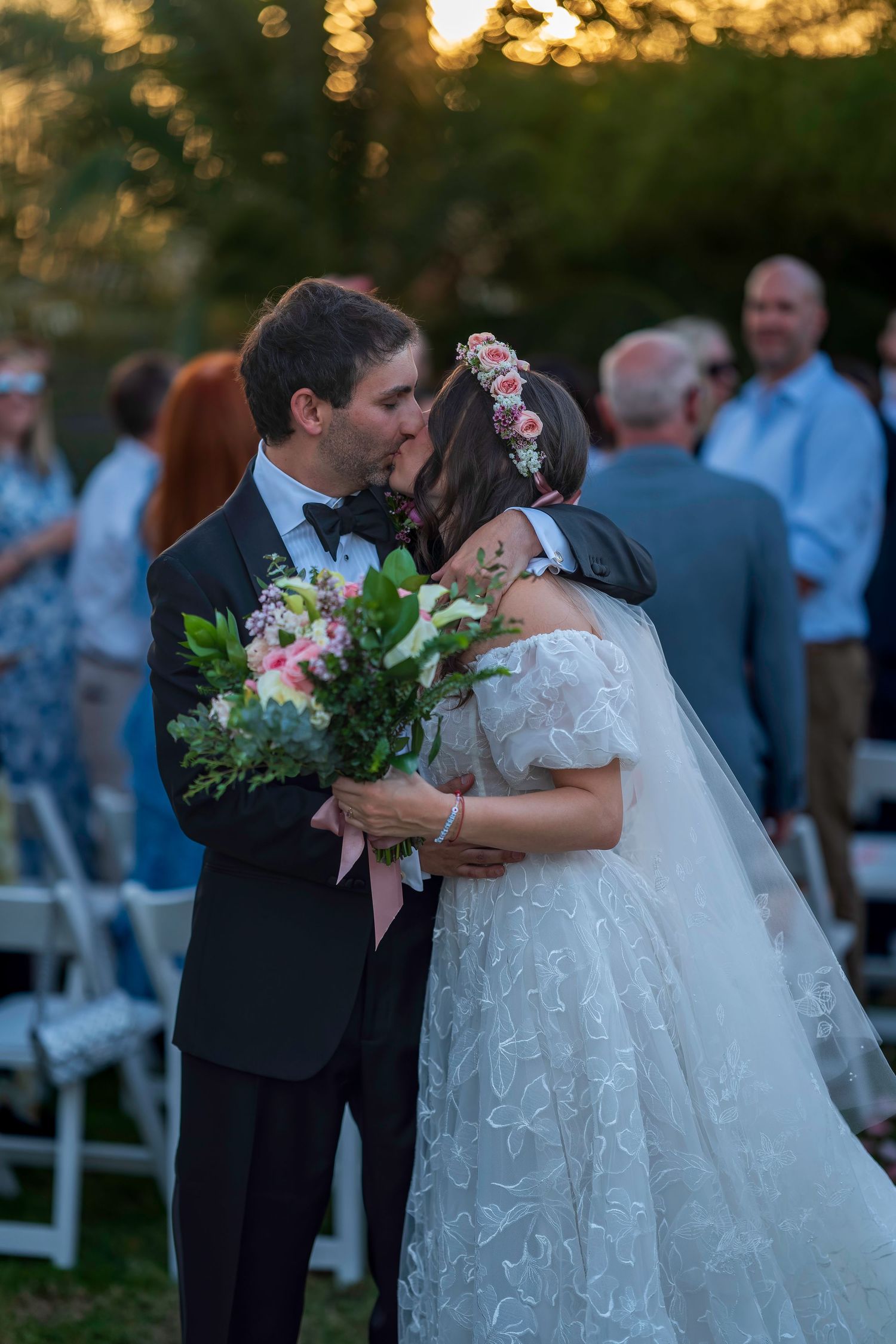 Bride and groom share first kiss at outdoor wedding ceremony, bride holding pink floral bouquet wearing lace gown and flower crown.