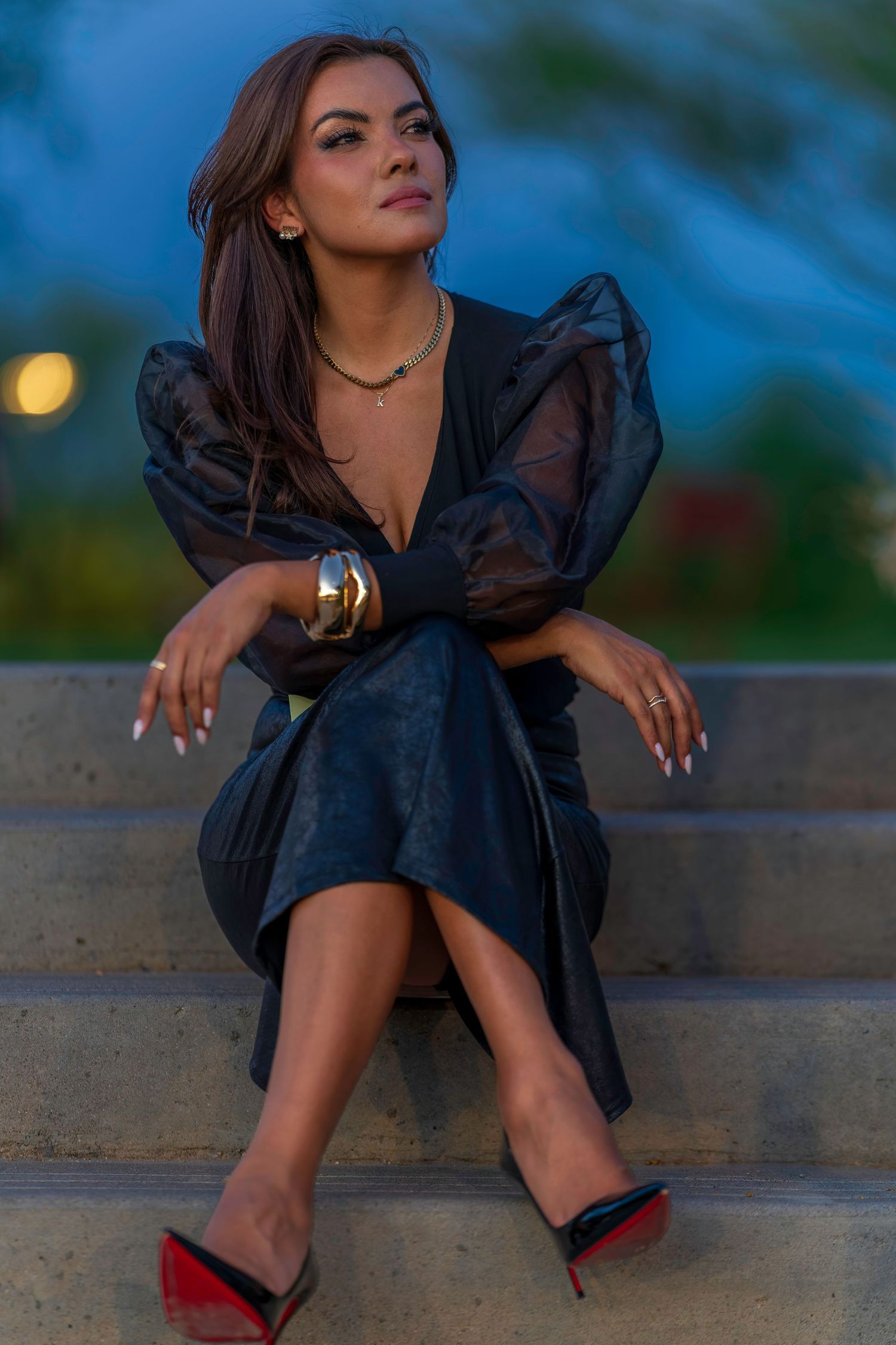Woman in black dress with sheer sleeves and gold bracelet sitting on steps wearing red heels at dusk.