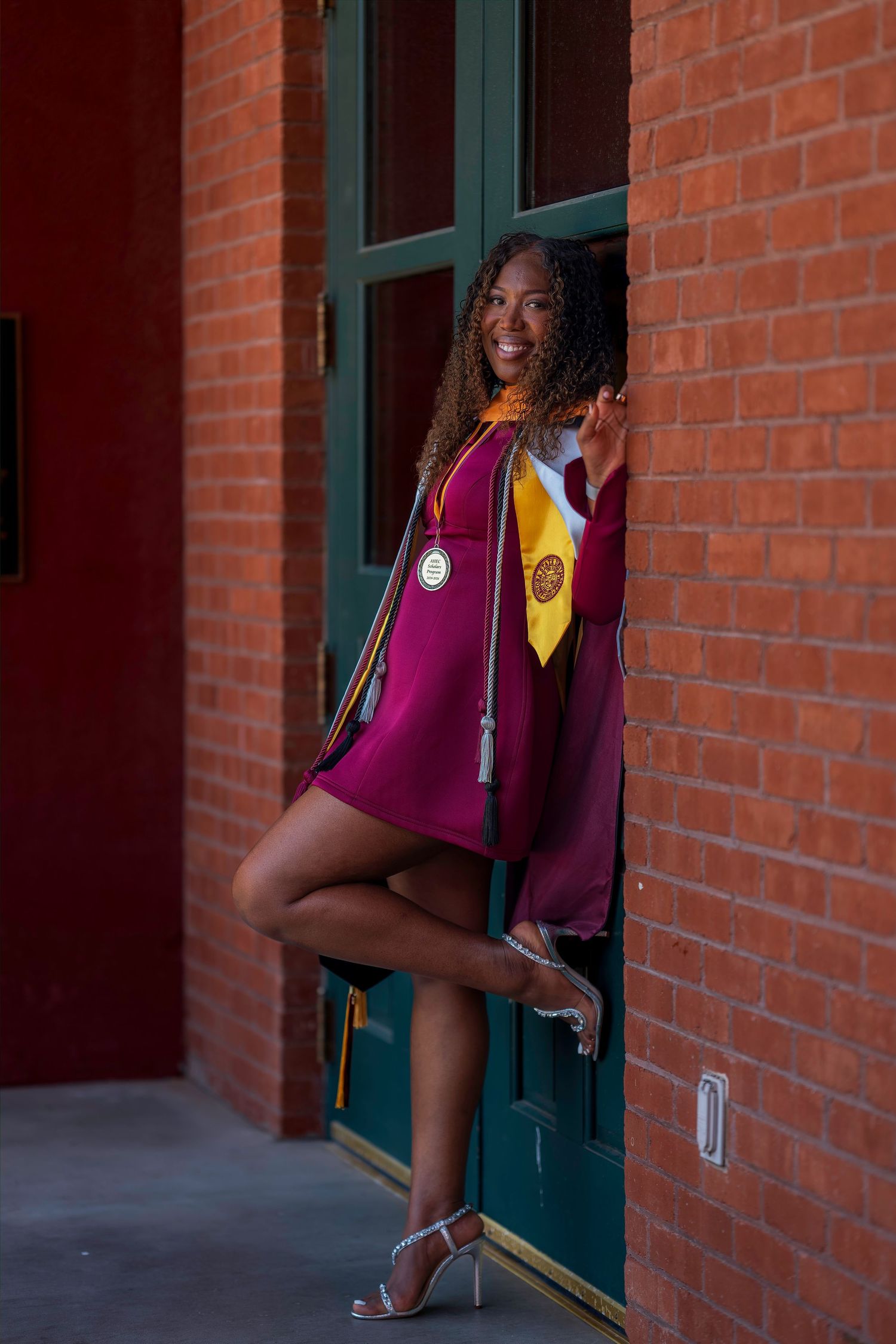 Young woman in maroon ASU graduation gown and stoles posing against brick wall with green door.