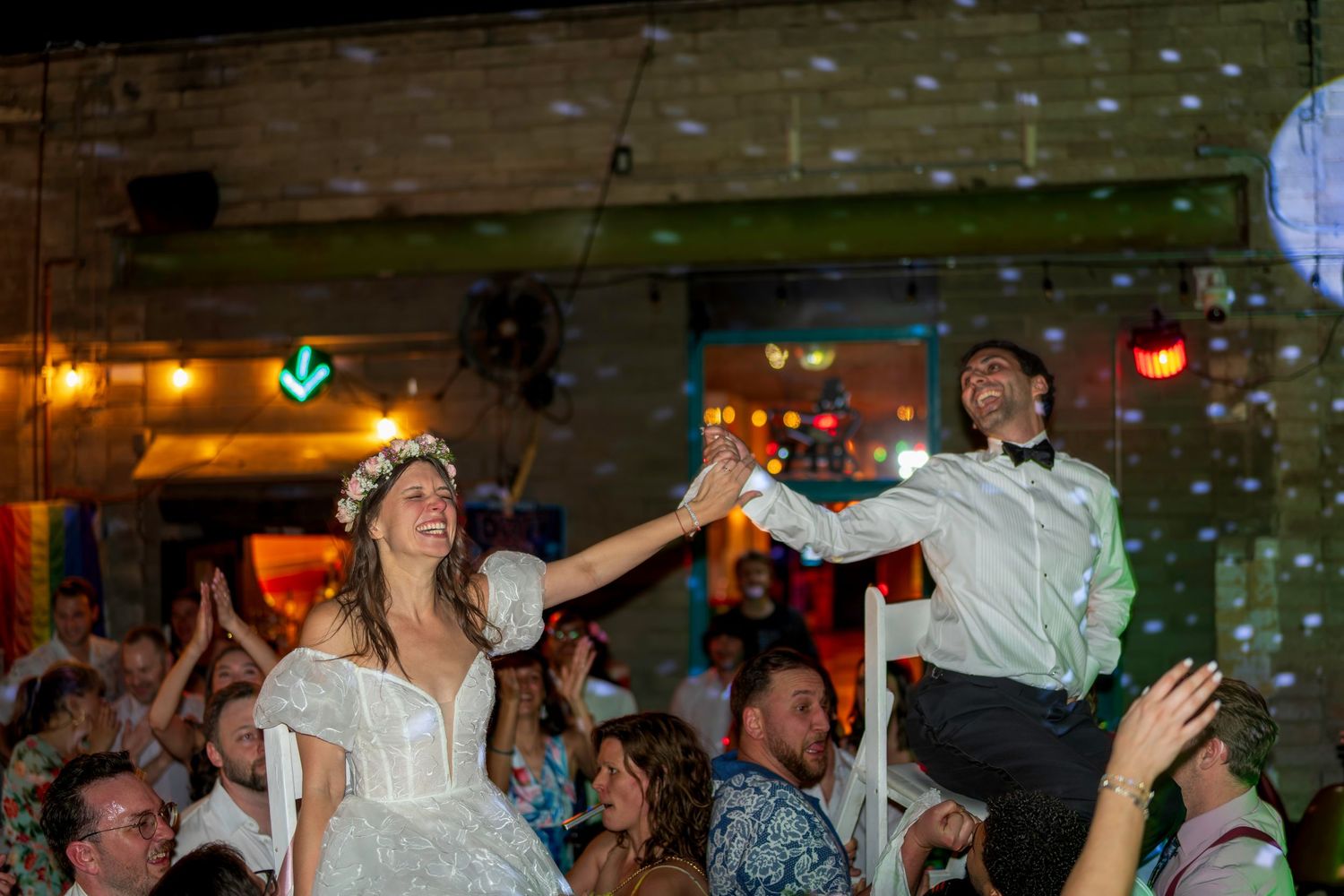 Bride and groom lifted on chairs during lively wedding reception dance celebration with festive lights.