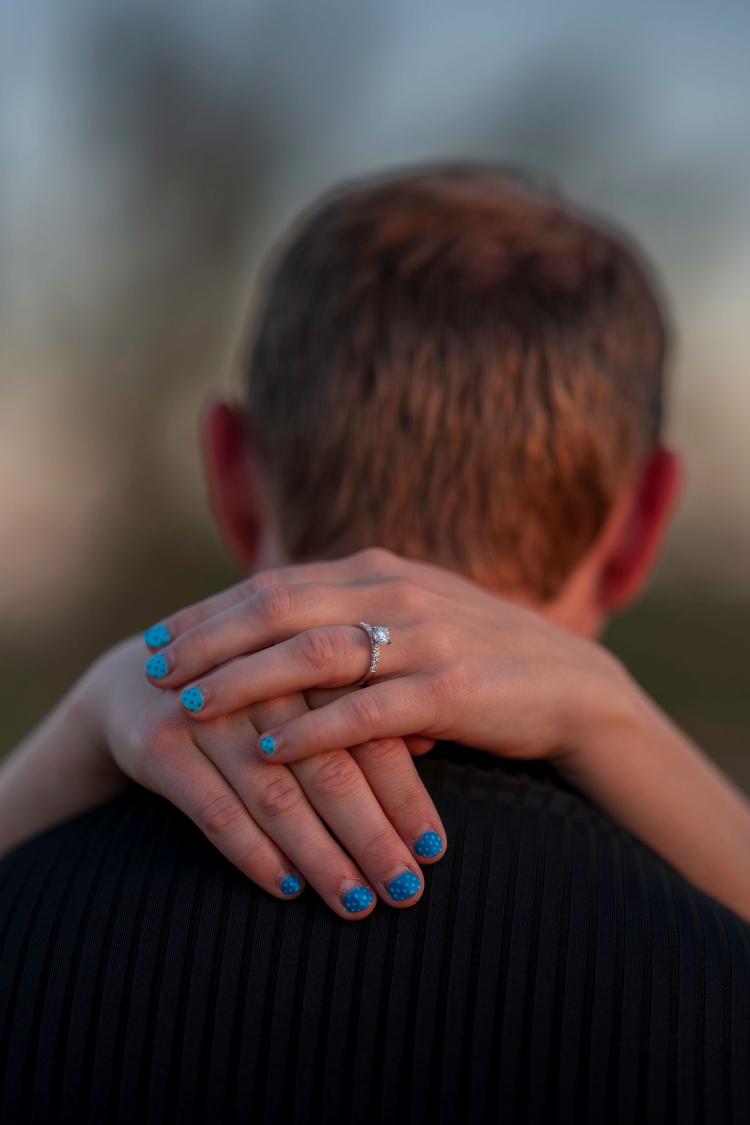 Close-up of a woman's hand with blue nails and engagement ring resting on a man's shoulder.
