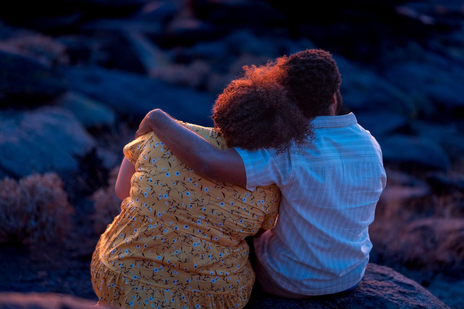Couple sitting together on rocks at dusk, woman in yellow floral dress leaning on mans shoulder.