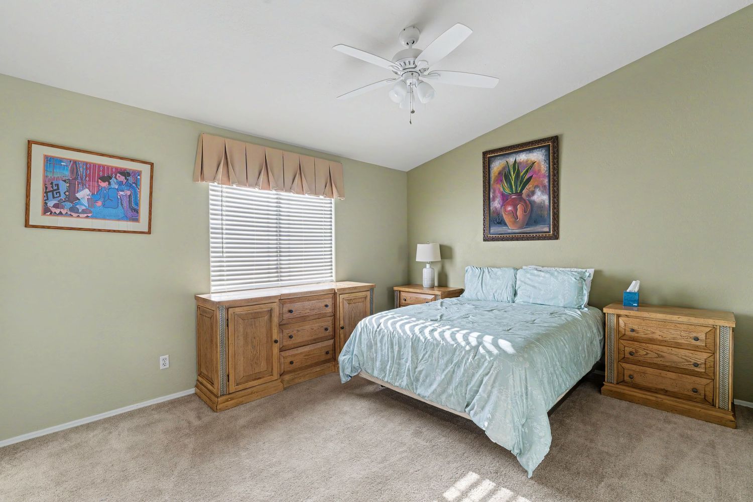 Spacious bedroom with light green walls, wooden dresser, queen bed with blue bedding, carpet flooring, and ceiling fan.