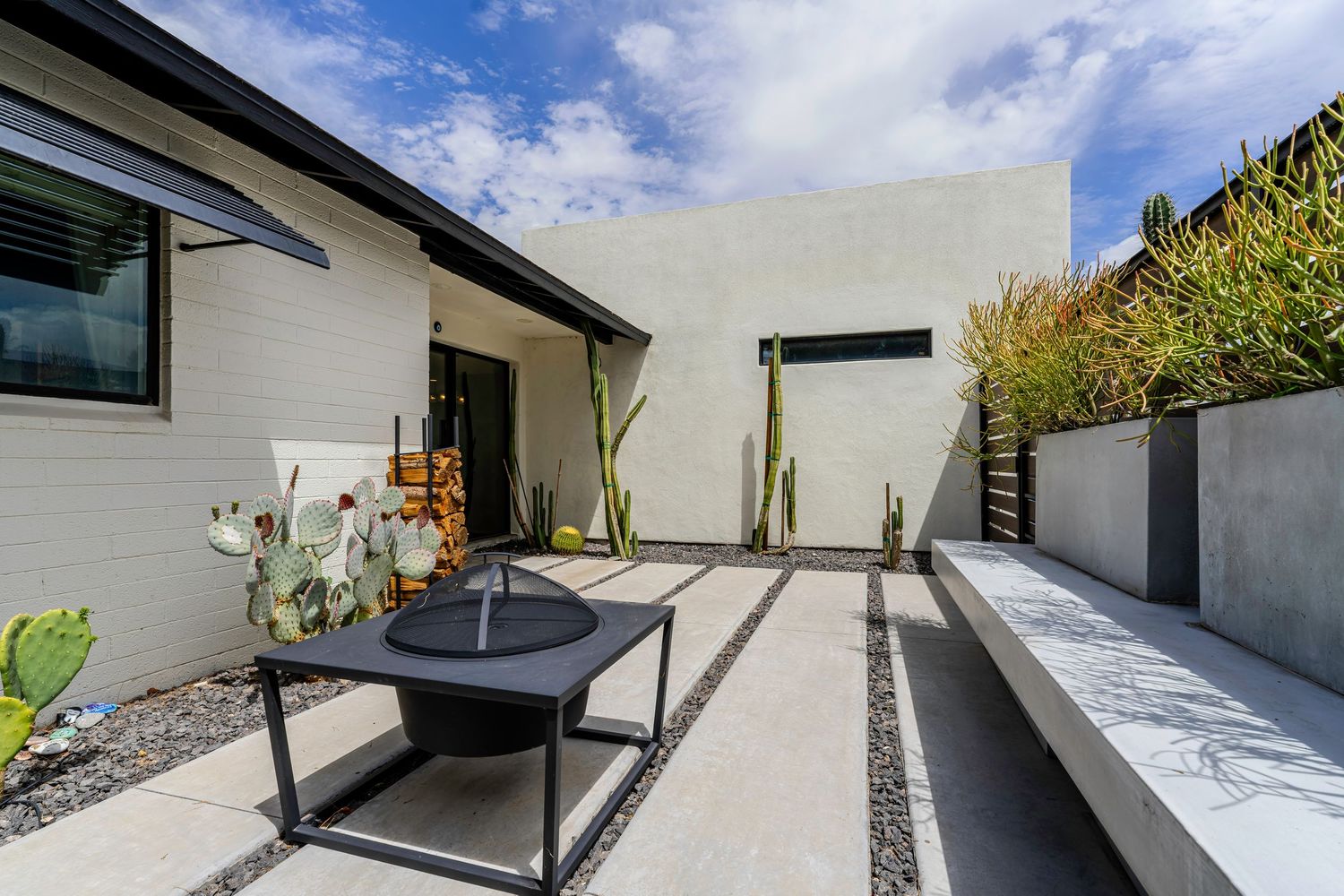 Modern home courtyard featuring white stucco walls, cacti, black fire pit table, and sleek concrete bench seating.