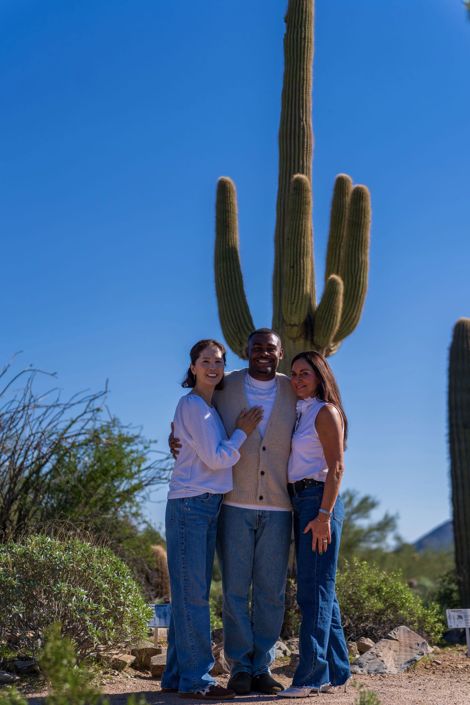 Three people pose together in front of a towering saguaro cactus in the desert.