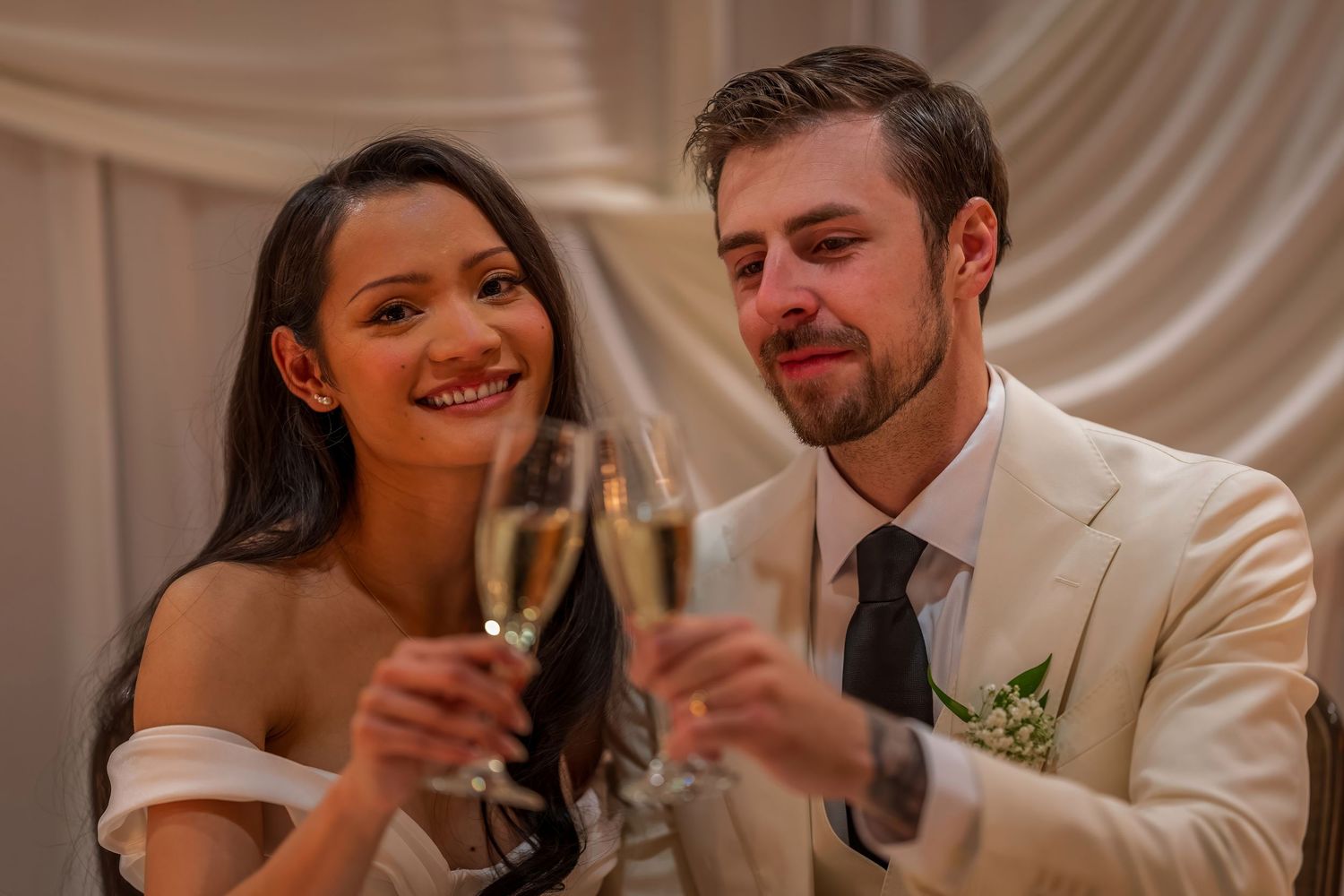Newlyweds share a romantic toast at their wedding reception with elegant white decor and draping in the background.