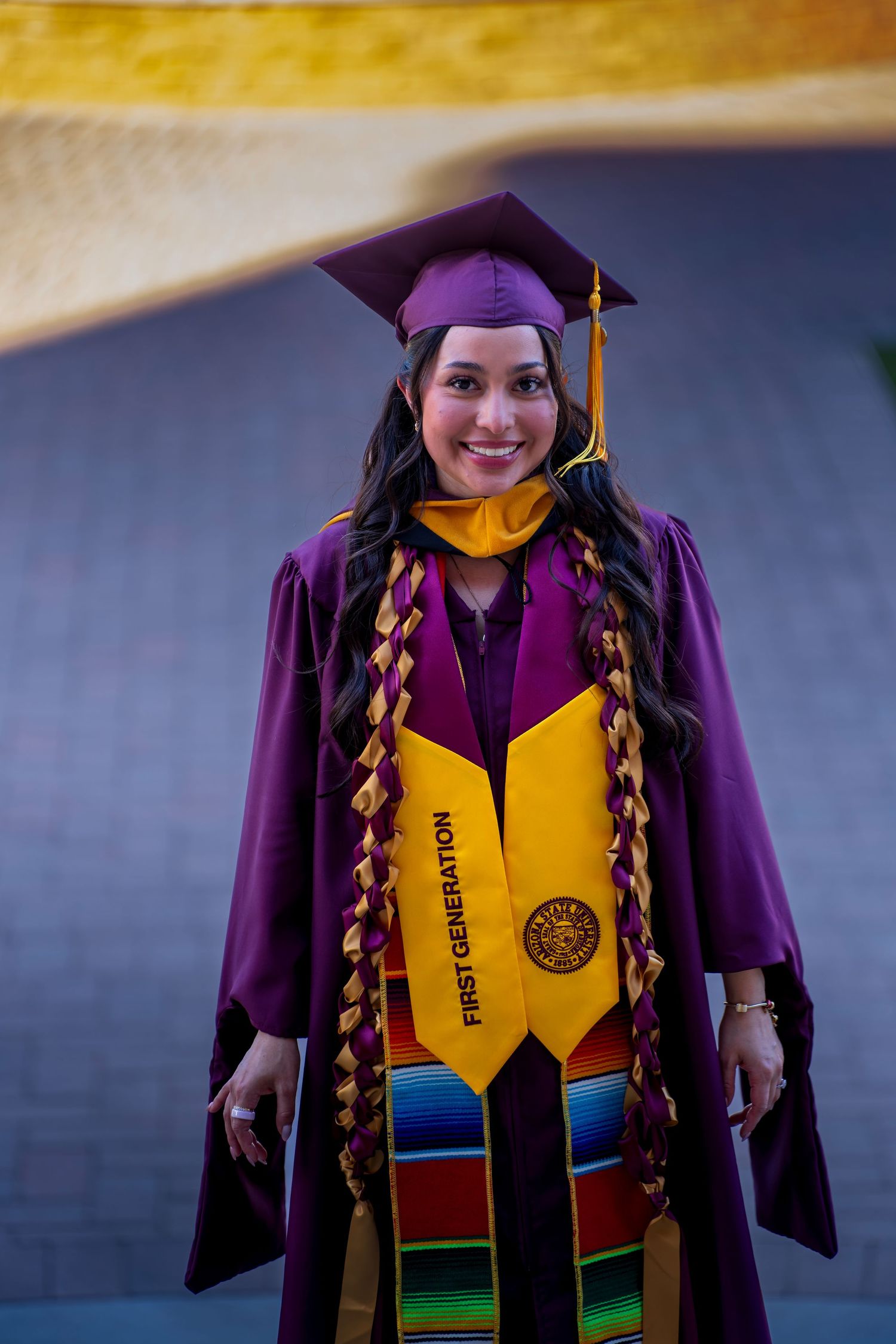 Graduate wearing purple academic regalia with yellow stole and colorful kente cloth stole during commencement ceremony.