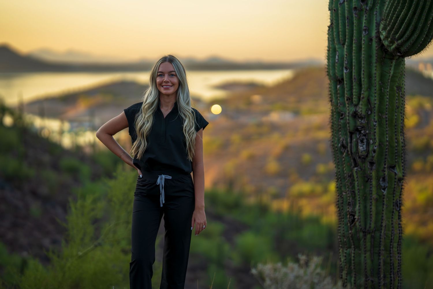 Healthcare professional in black scrubs poses against desert sunset with saguaro cactus and mountain backdrop.