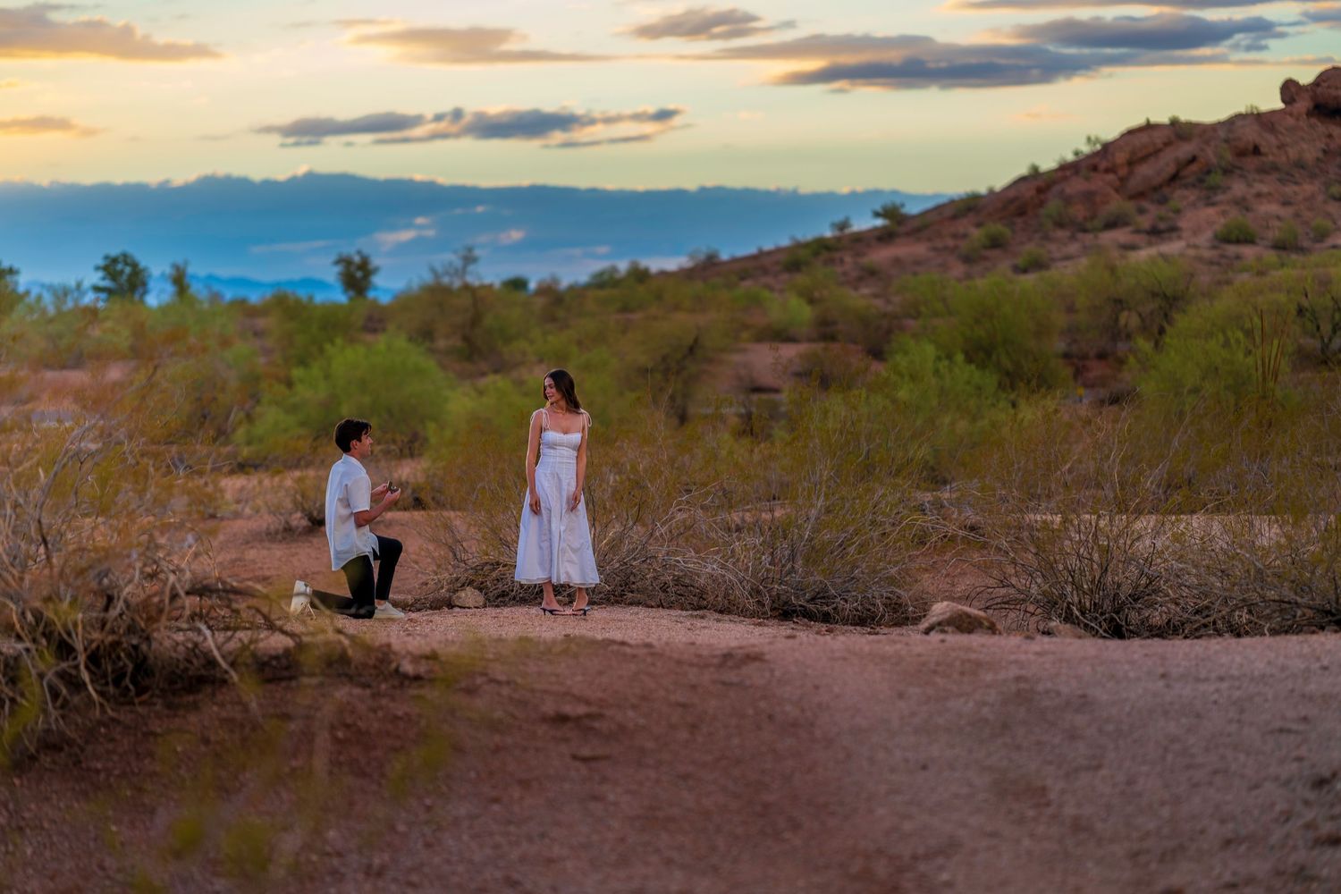 A couple shares their engagement moment on a desert trail at sunset.