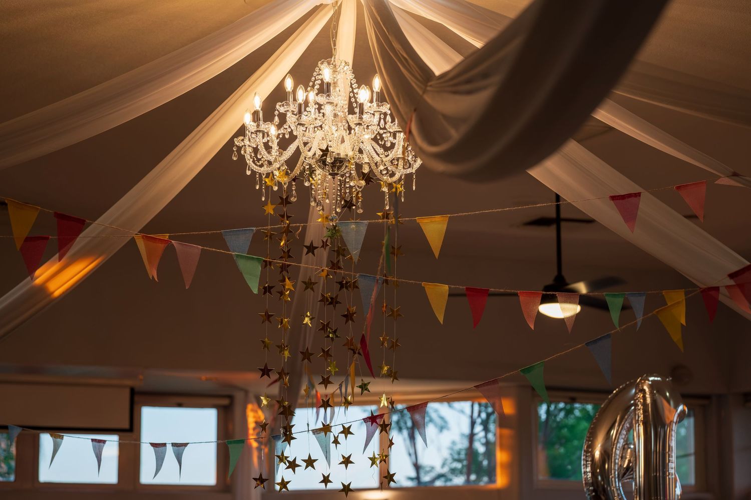 An elegant crystal chandelier hangs beneath draped fabric and colorful bunting flags at an event venue.