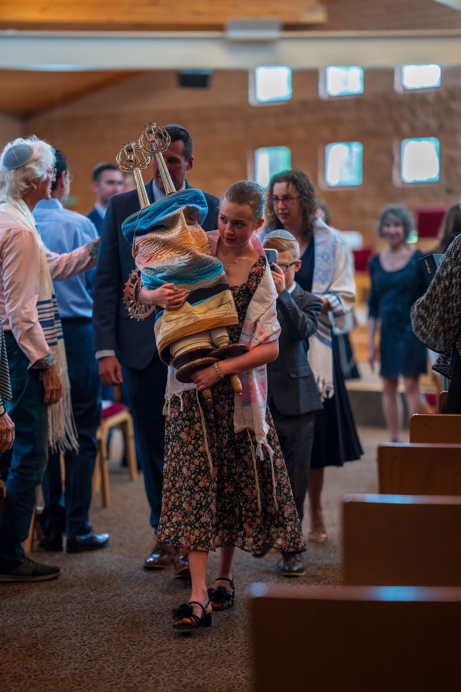 Young girl carries Torah scrolls during Jewish synagogue ceremony, wrapped in tallit, with congregation gathered in background.