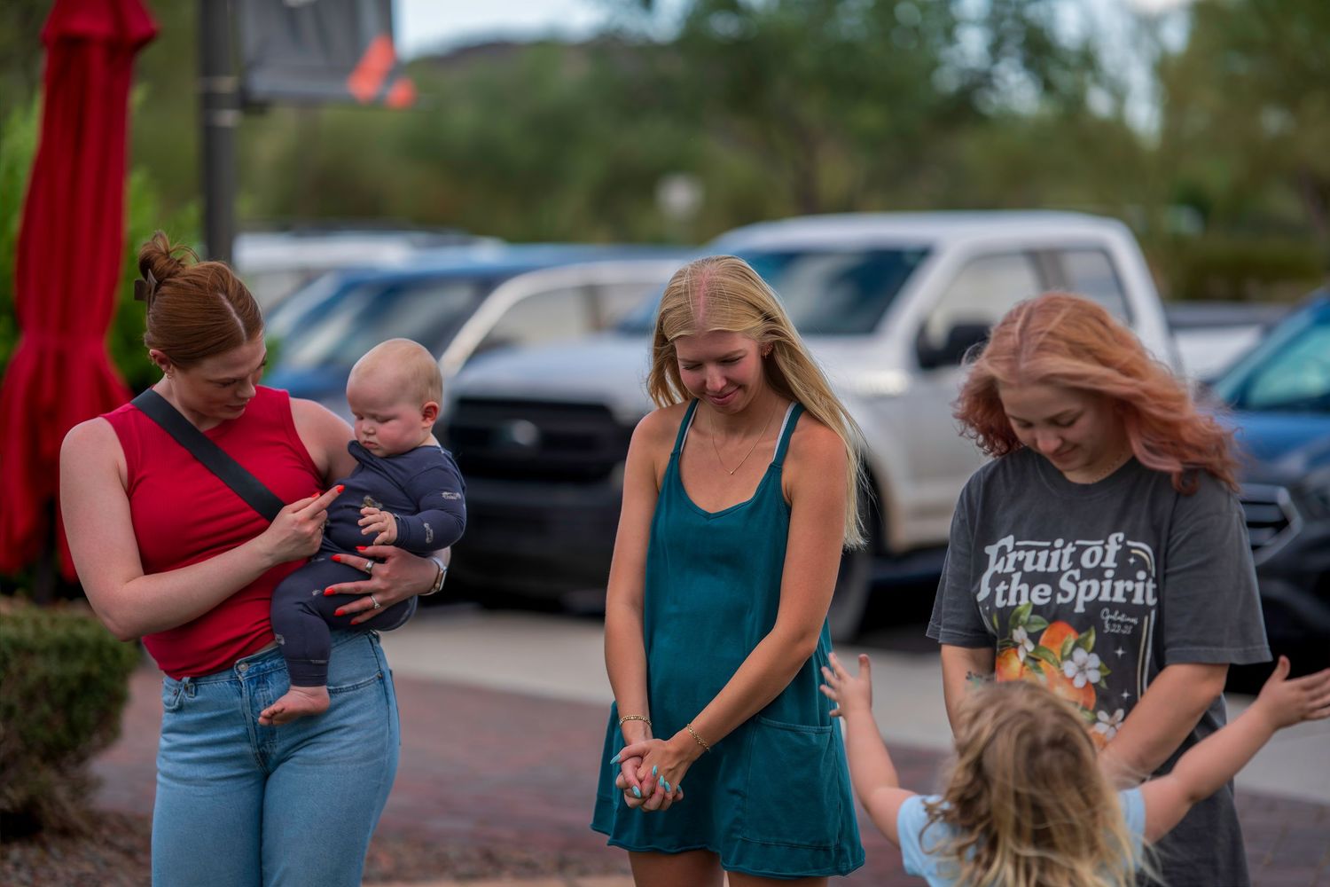 Women and children gather outdoors in a parking lot, one holding a baby, smiling and socializing on a warm evening.