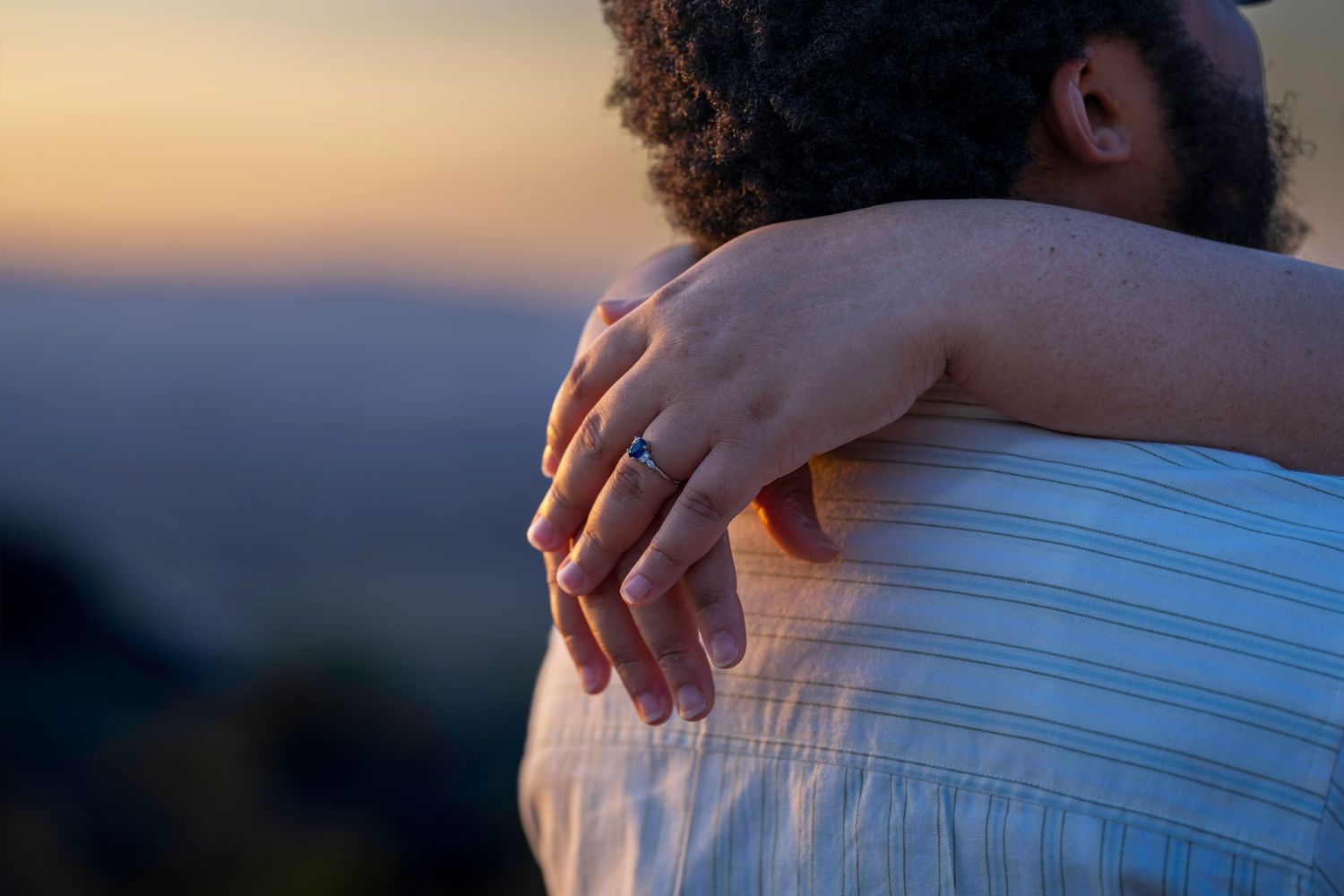 Couple embracing at sunset, woman's hand with engagement ring resting on man's shoulder.