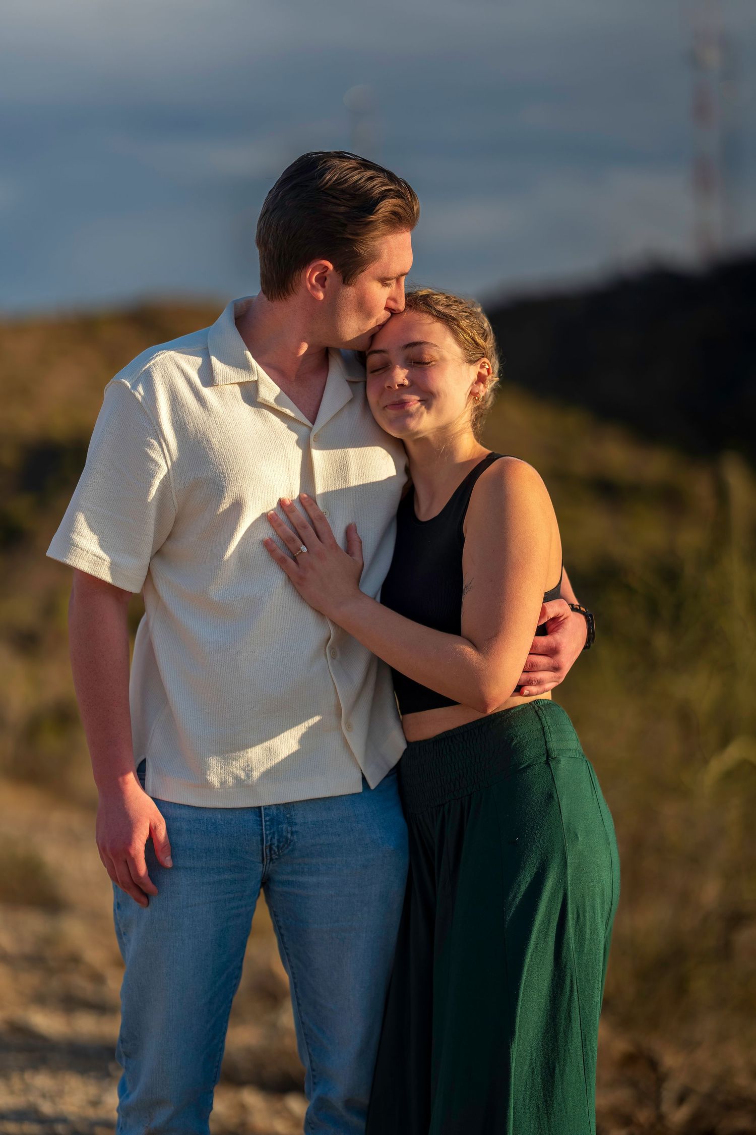 Romantic couple sharing a tender moment at sunset, man kissing womans temple while she smiles peacefully.