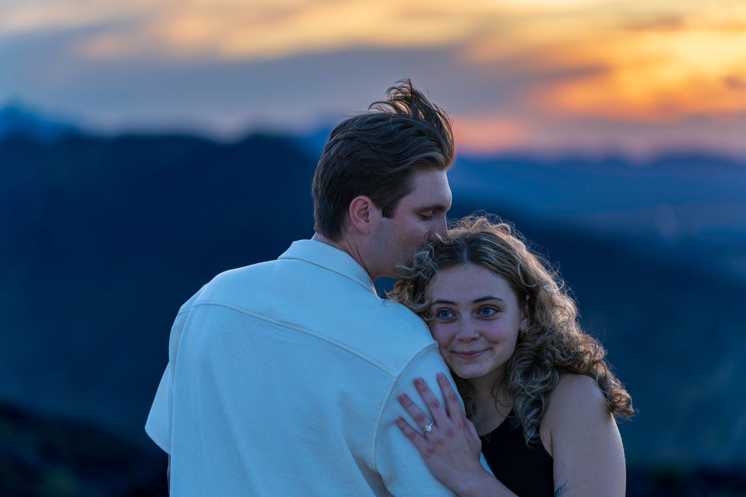 A couple shares a tender embrace at sunset with colorful mountain views in the background.