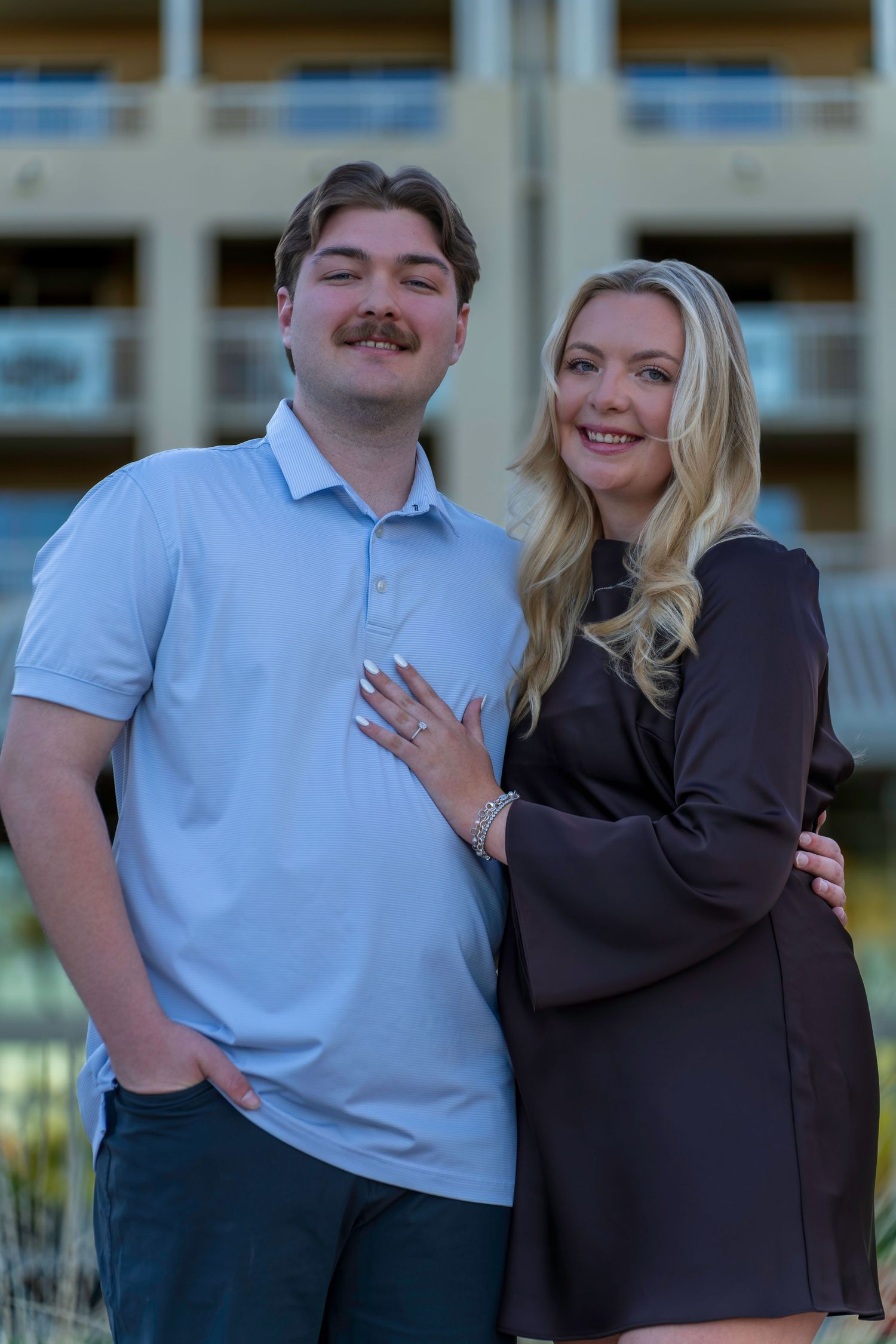 Young couple posing outdoors, woman showing engagement ring, man in blue polo shirt, building in background.