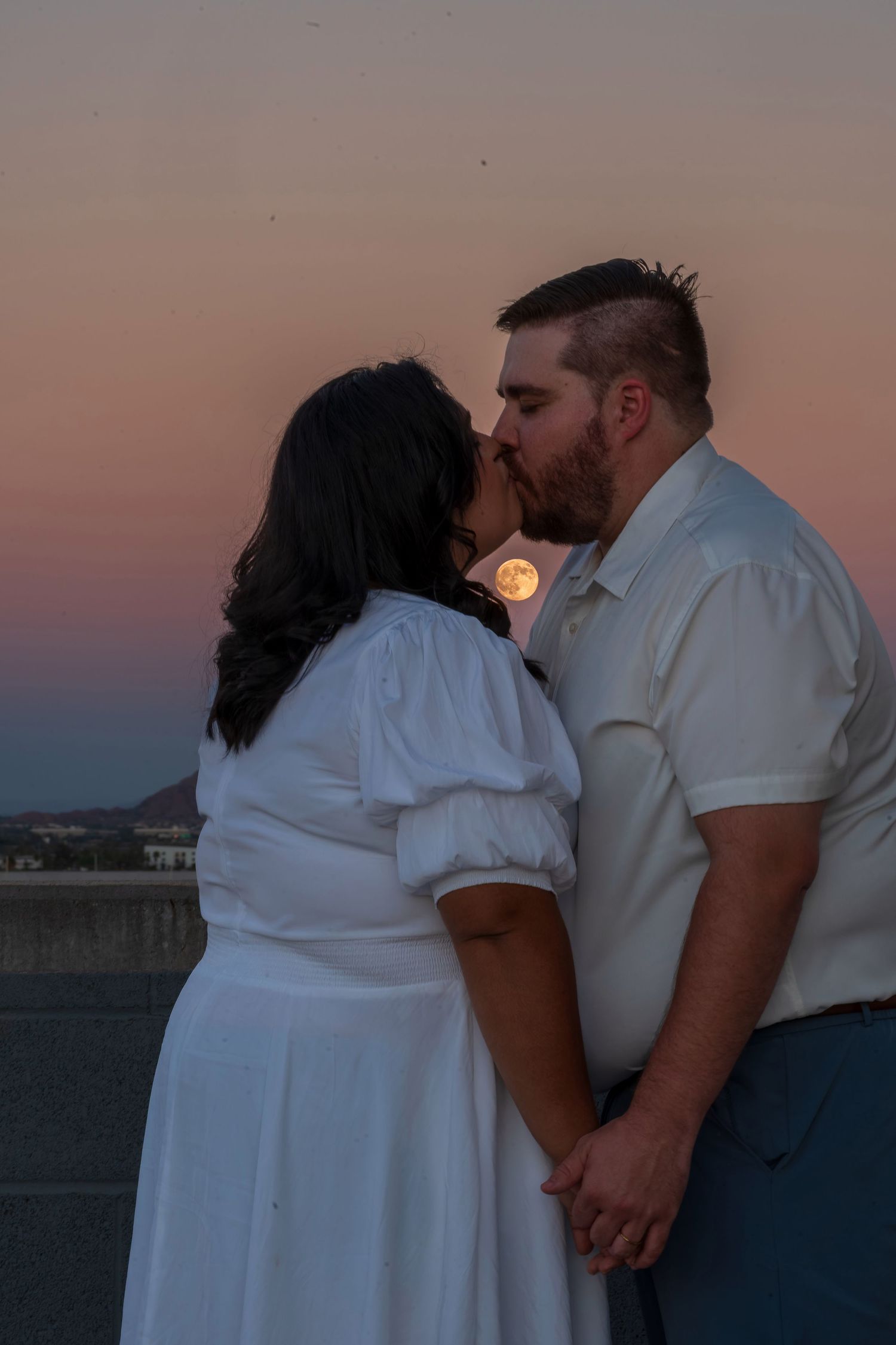Couple kissing at sunset with full moon rising behind them, woman in white dress holding hands with man in white shirt.