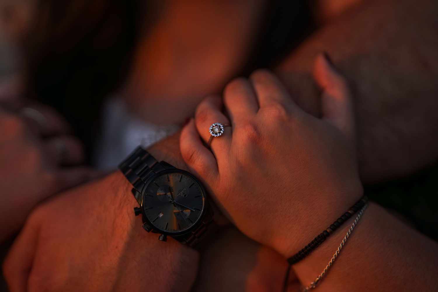 Close-up of couples hands touching, featuring diamond engagement ring and black watch at sunset.