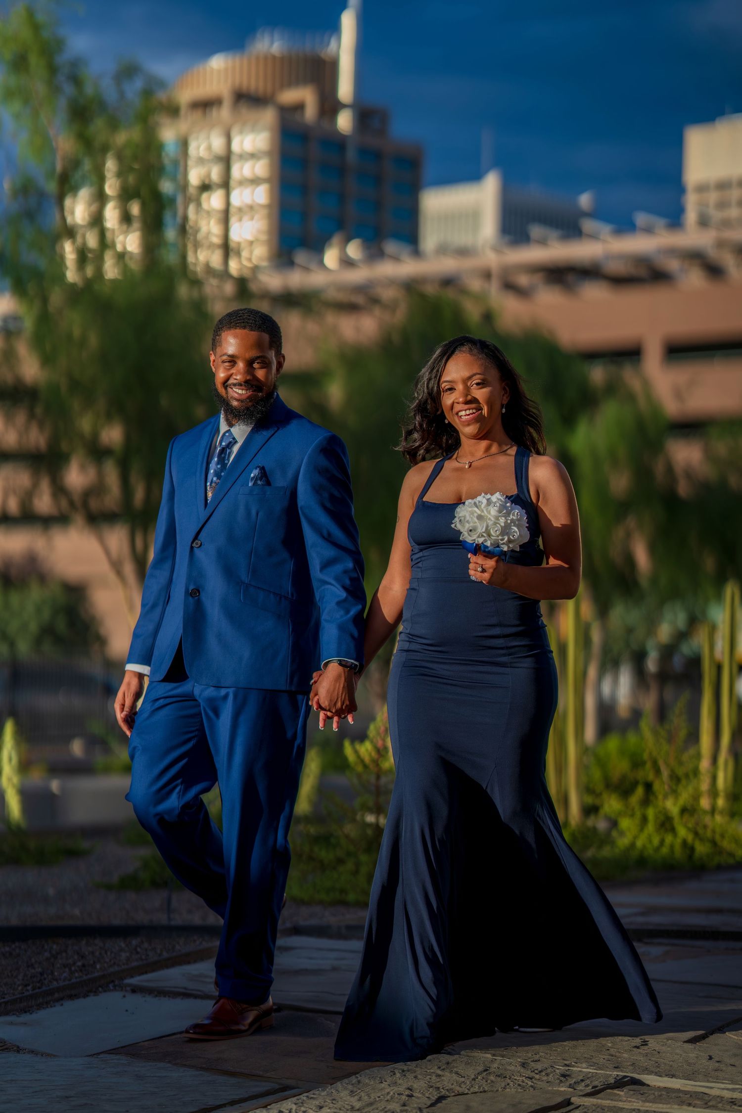 Couple in coordinating blue wedding attire walking hand-in-hand outdoors with city buildings in background.