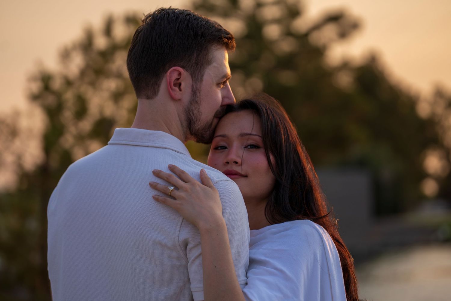 Couple embracing tenderly at golden sunset, woman in white dress rests head on mans shoulder.