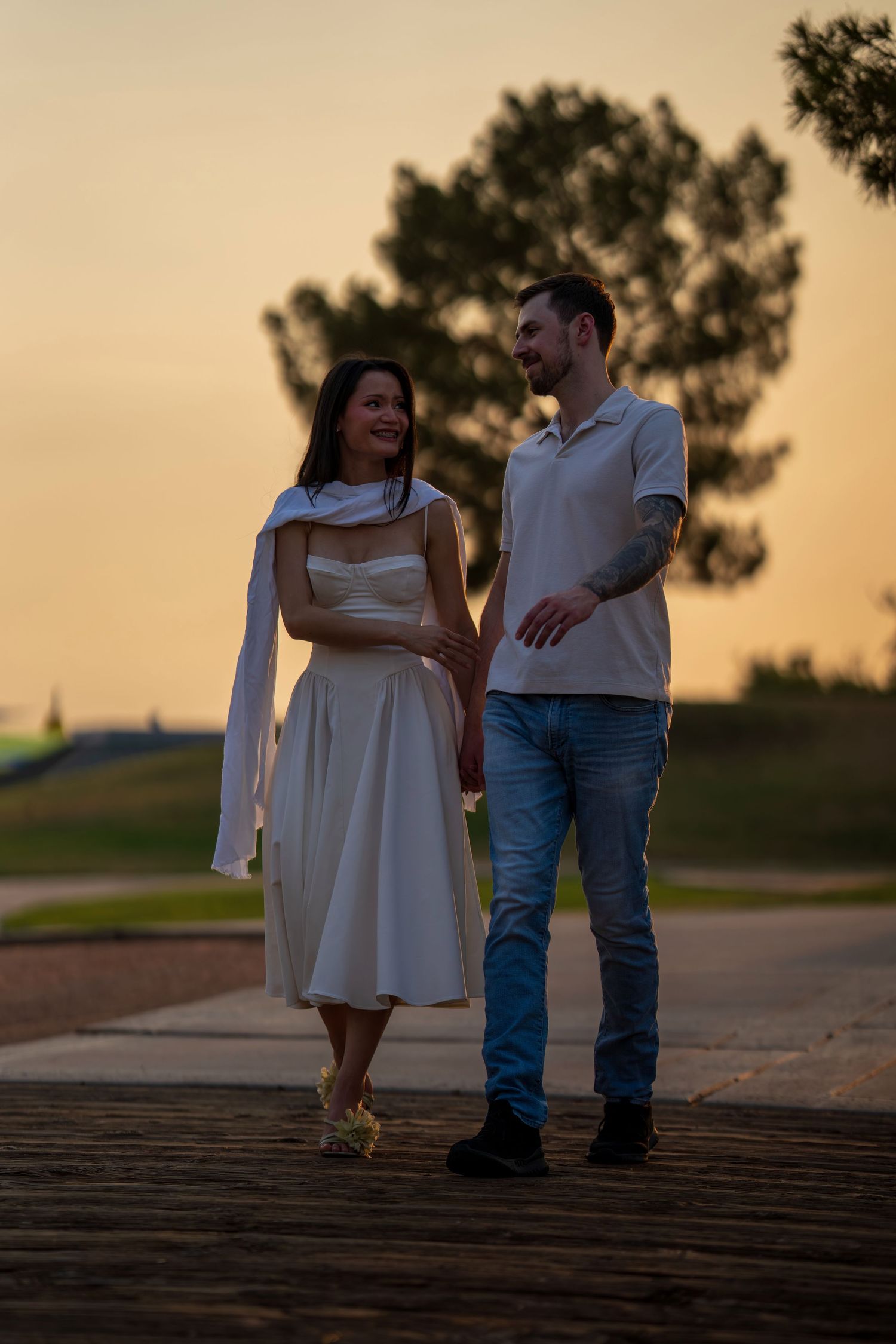 Couple walking together on wooden path at sunset, woman in white midi dress and man in jeans.