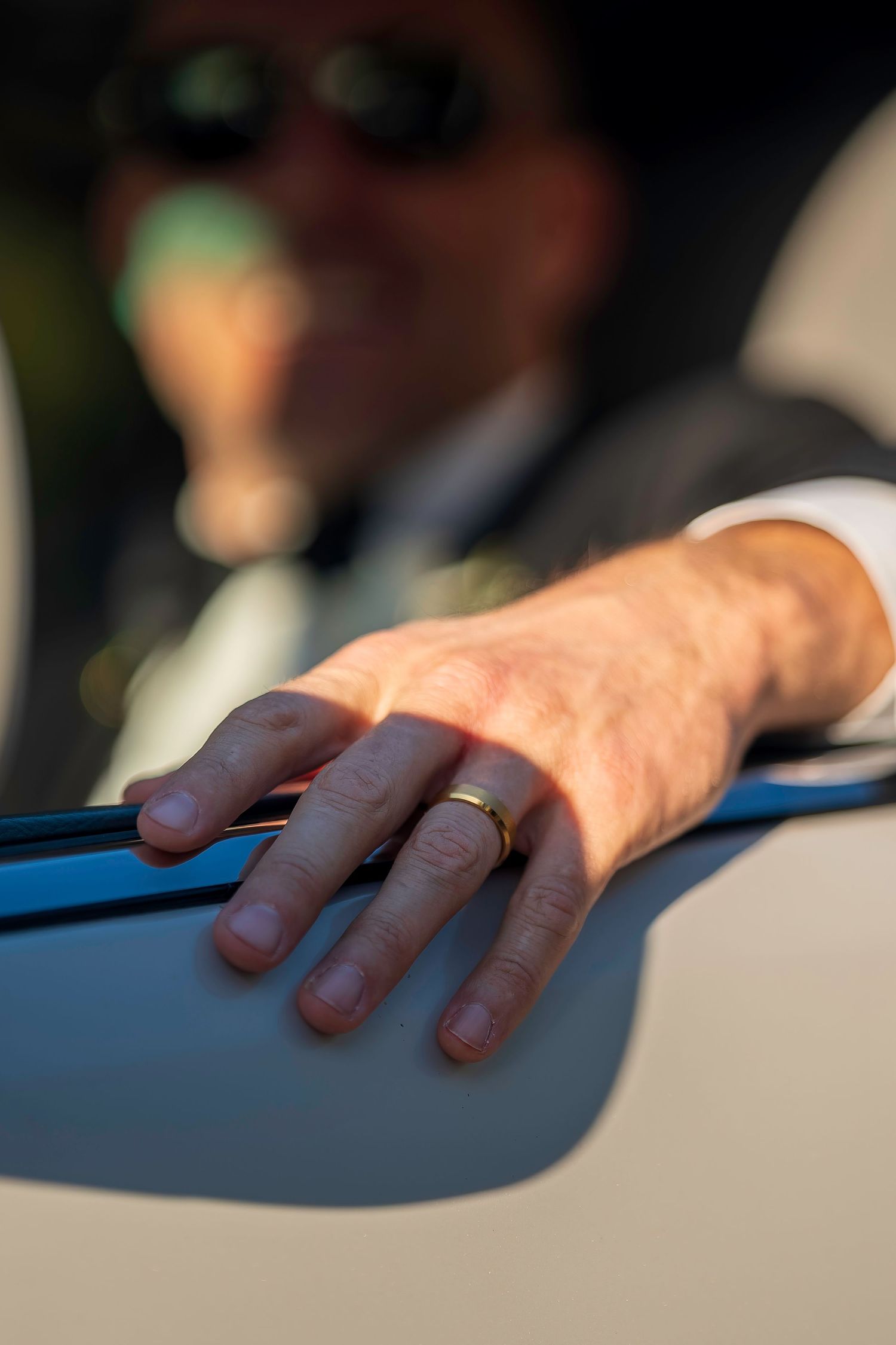 Close-up of groom's hand with gold wedding band resting on car door after ceremony.