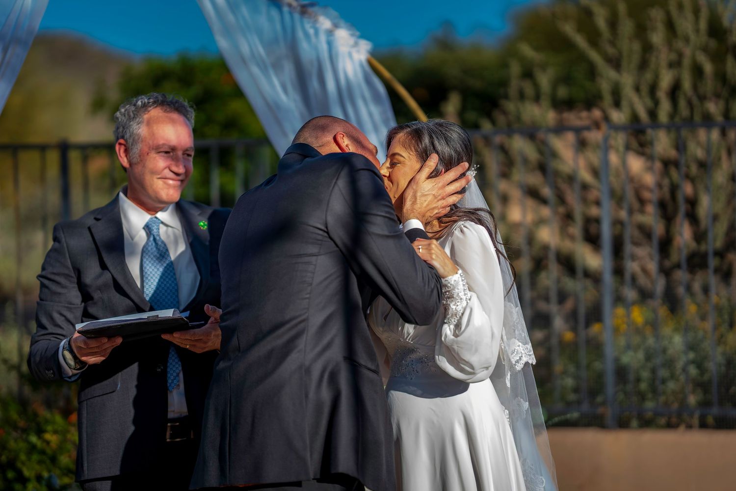 Bride and groom share first kiss at outdoor wedding ceremony while officiant smiles nearby.