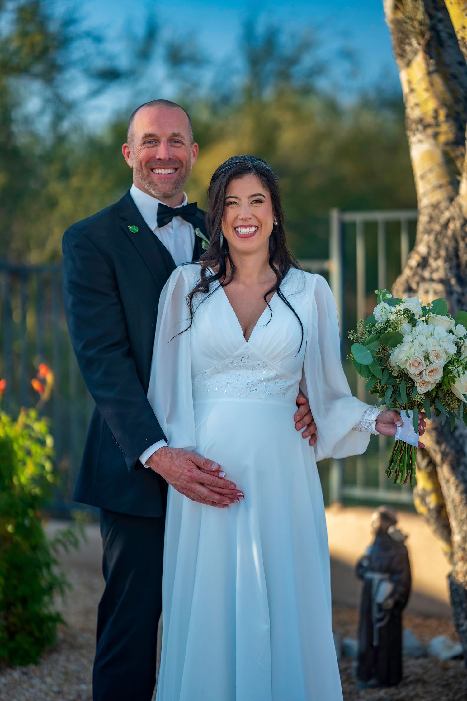 Happy bride and groom posing together outdoors, bride holding white floral bouquet in desert garden.