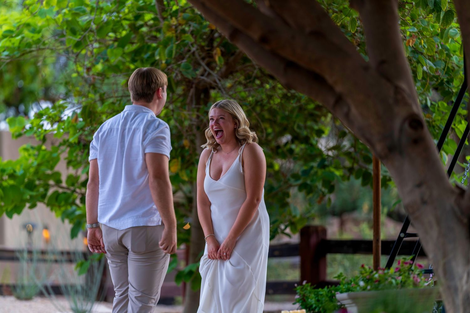 Couple in casual elegant attire sharing a romantic moment under a tree during an outdoor proposal.