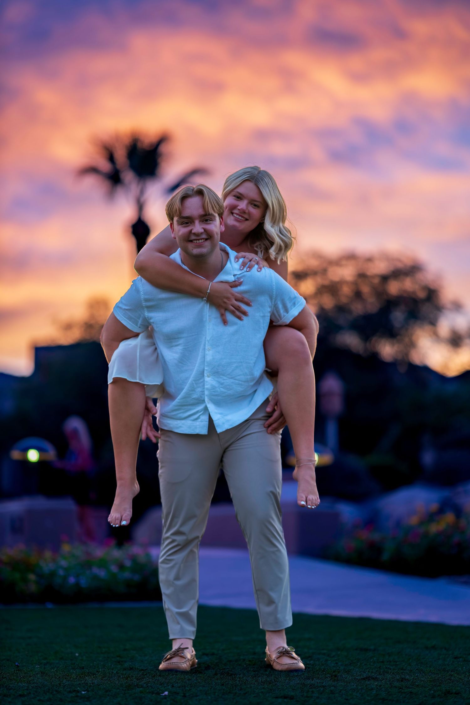 A joyful couple poses at sunset, man giving woman a piggyback ride against a vibrant purple and orange sky.