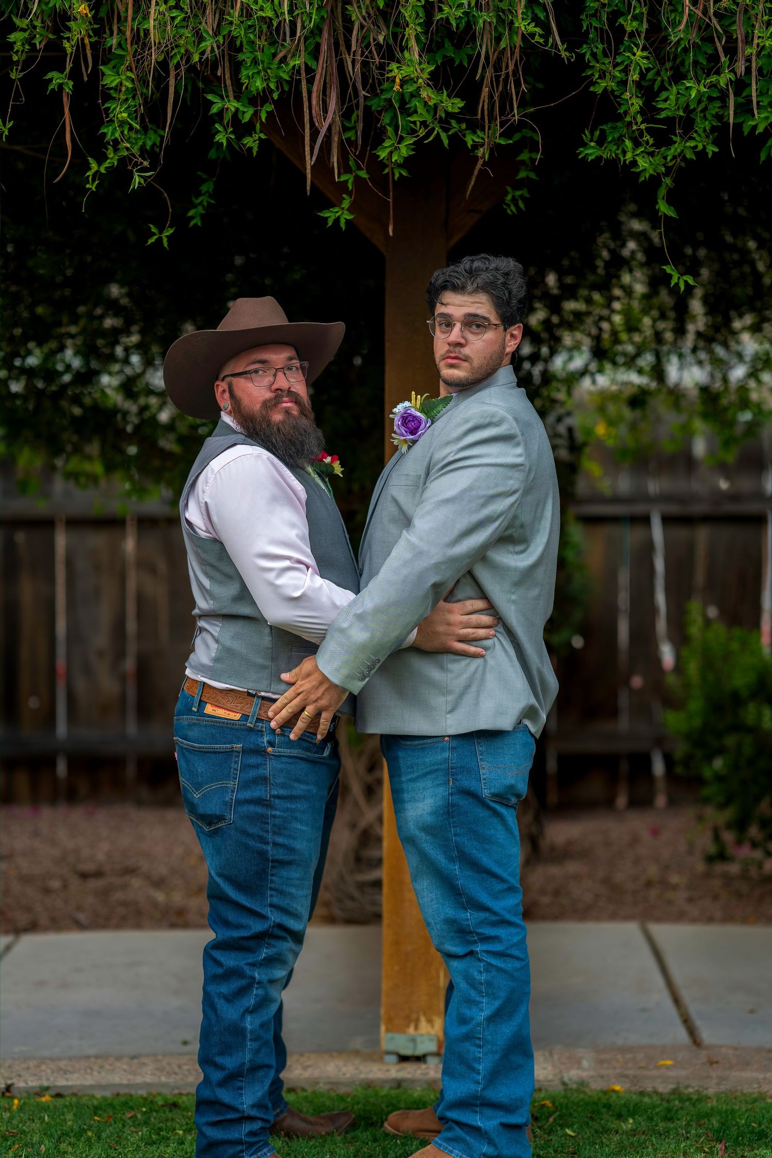 Two groomsmen in jeans and boutonnieres pose back-to-back under a vine-covered arbor outdoors.