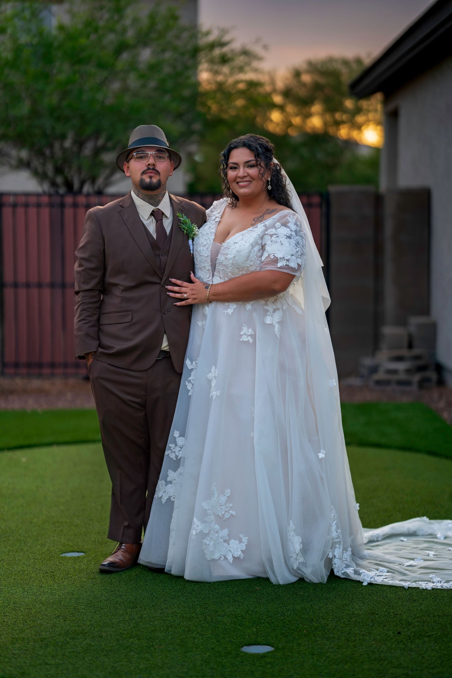 Bride in white floral lace gown poses with groom in brown suit on green lawn at sunset wedding.