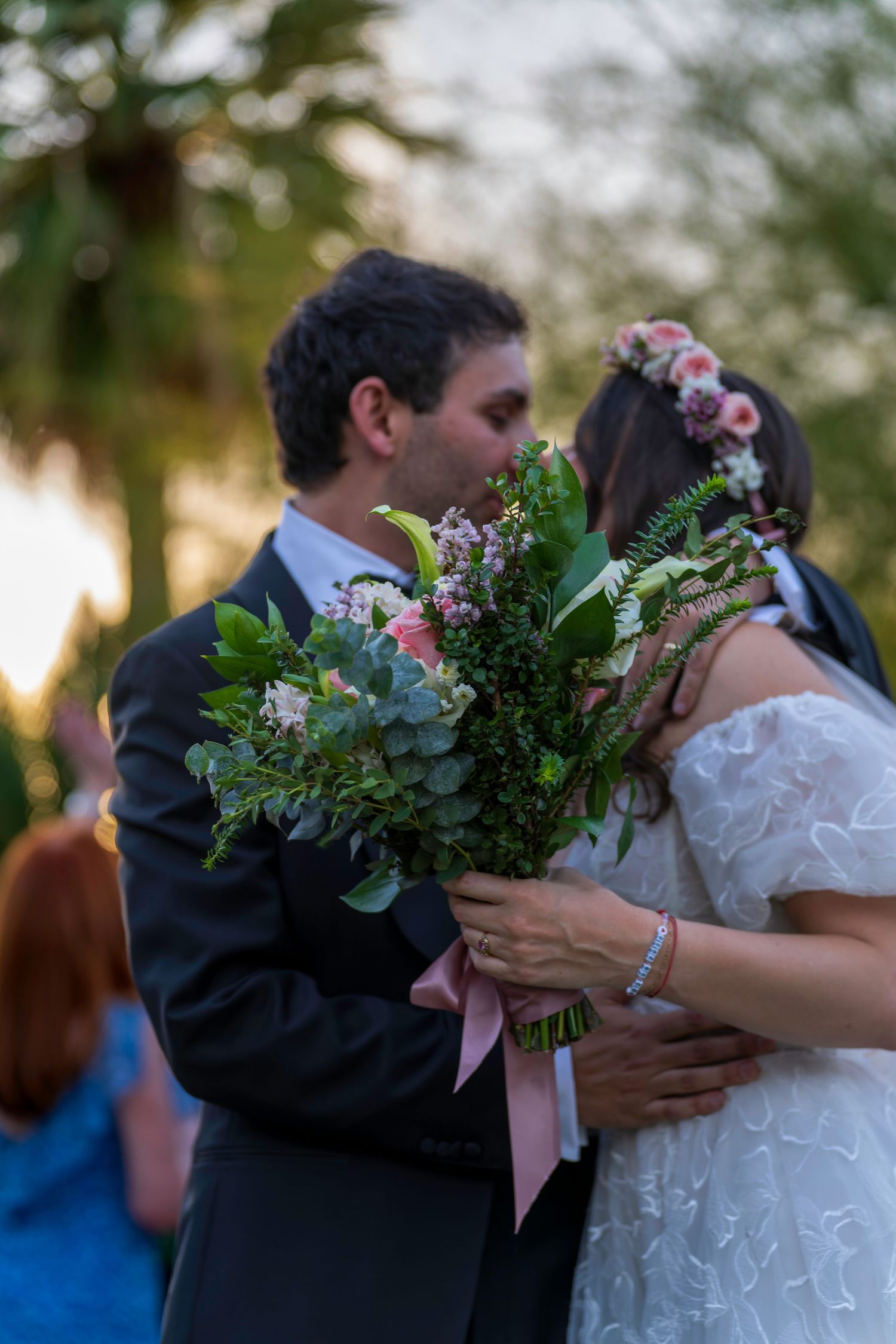 A bride and groom share a kiss behind a lush green and pink floral bouquet during their outdoor wedding ceremony.