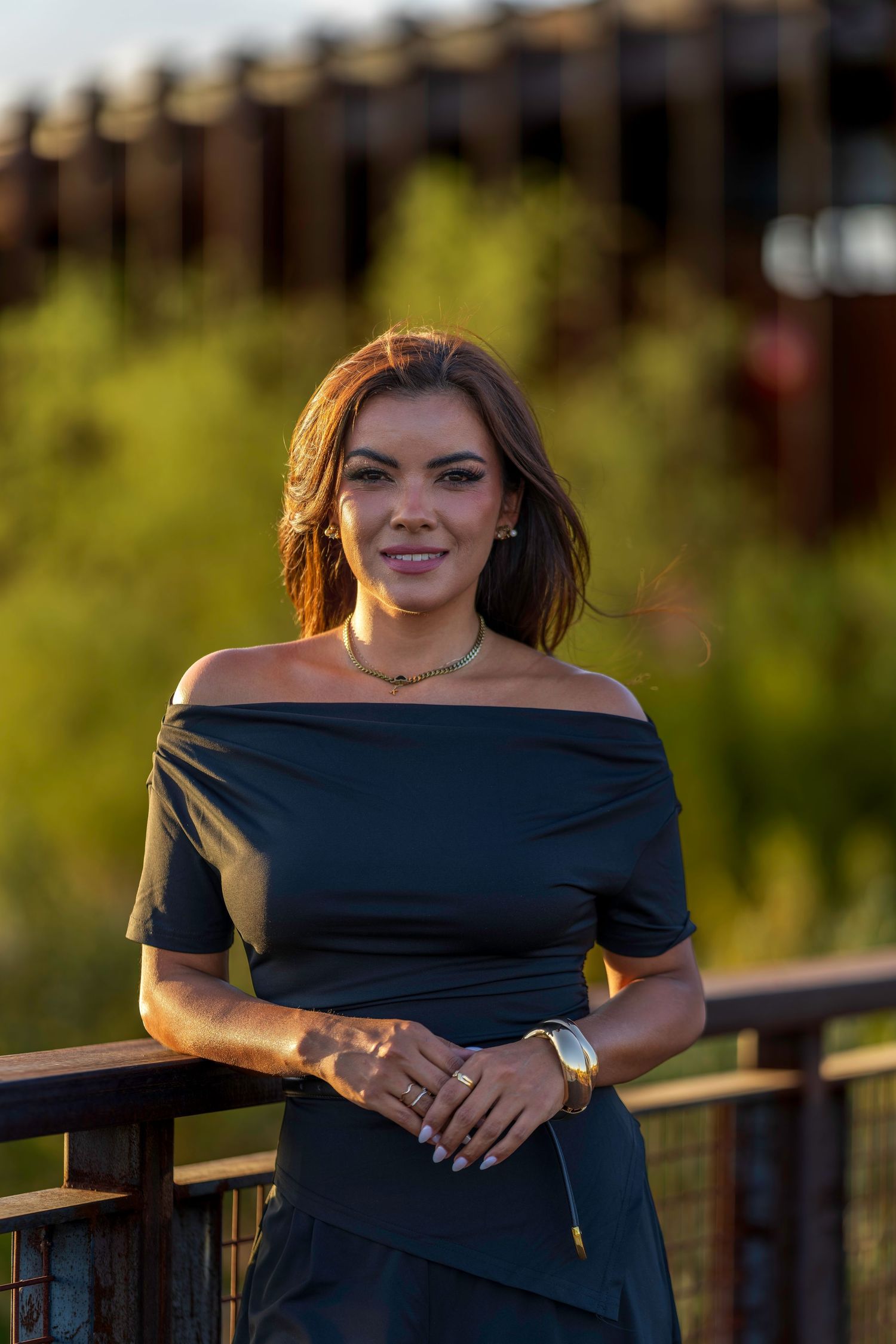 Woman in black off-shoulder dress smiling on a wooden deck with lush green background in golden hour light.