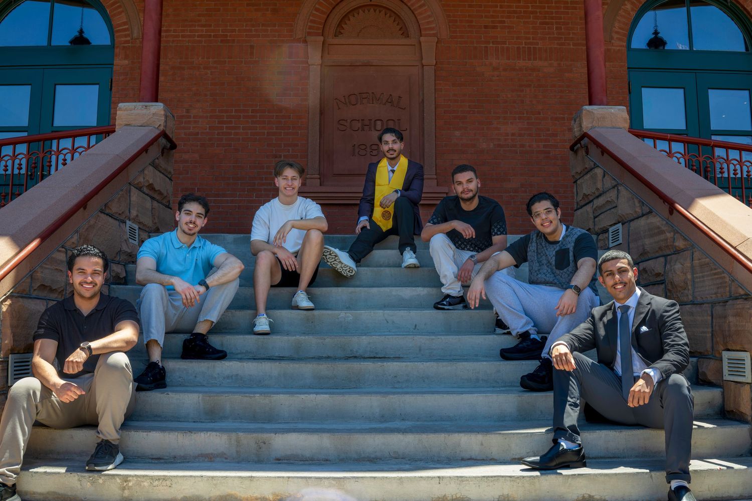 Seven young men sitting on brick building steps outdoors on a sunny day.