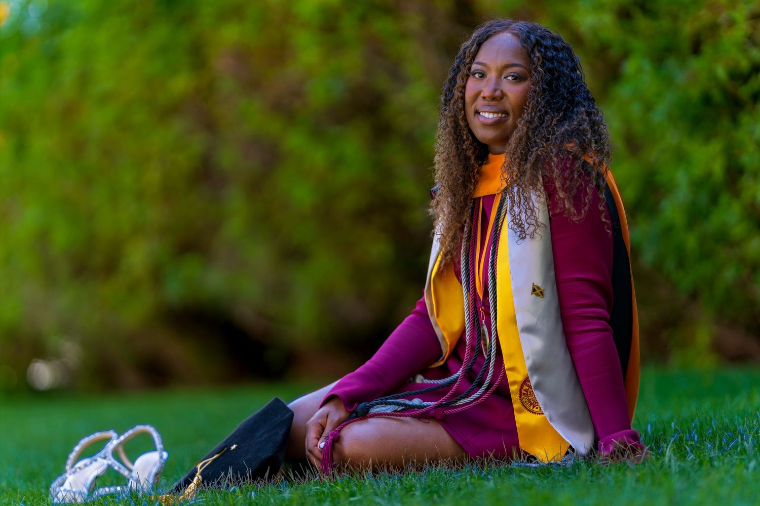 Smiling graduate in maroon and gold regalia sitting on green grass outdoors for a graduation portrait photo.